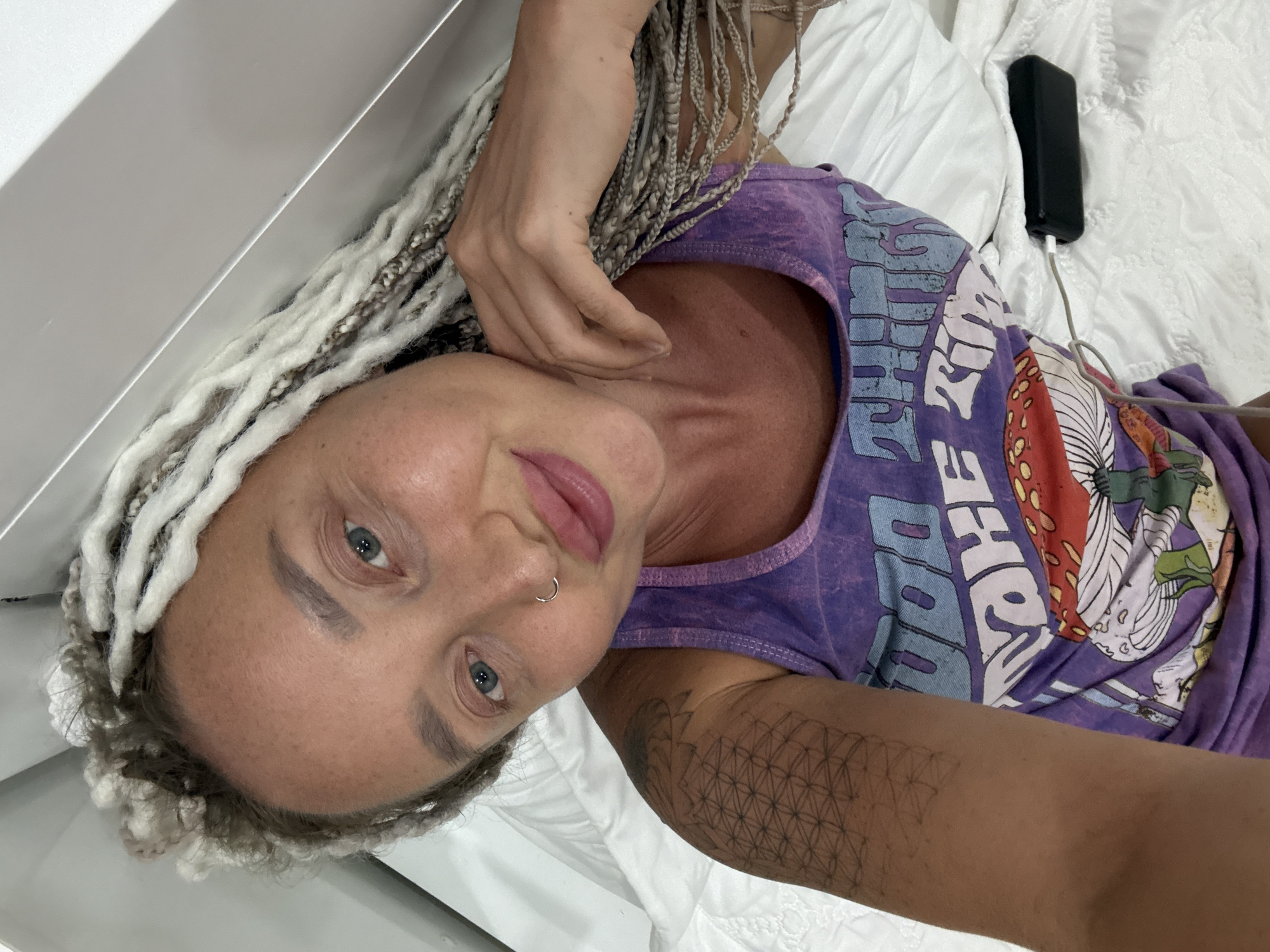 A woman with blonde braided hair lies on a white bed. She is wearing a purple tank top with a graphic print of mushrooms and text that is difficult to read. Her left hand is resting on her neck and cheek. A black power bank is visible on the bed to her right, plugged into a white cable. The woman has a nose ring and her eyes are open, looking towards the camera. The setting appears to be indoors, possibly a bedroom, with a white, textured bedspread. The lighting suggests daytime.