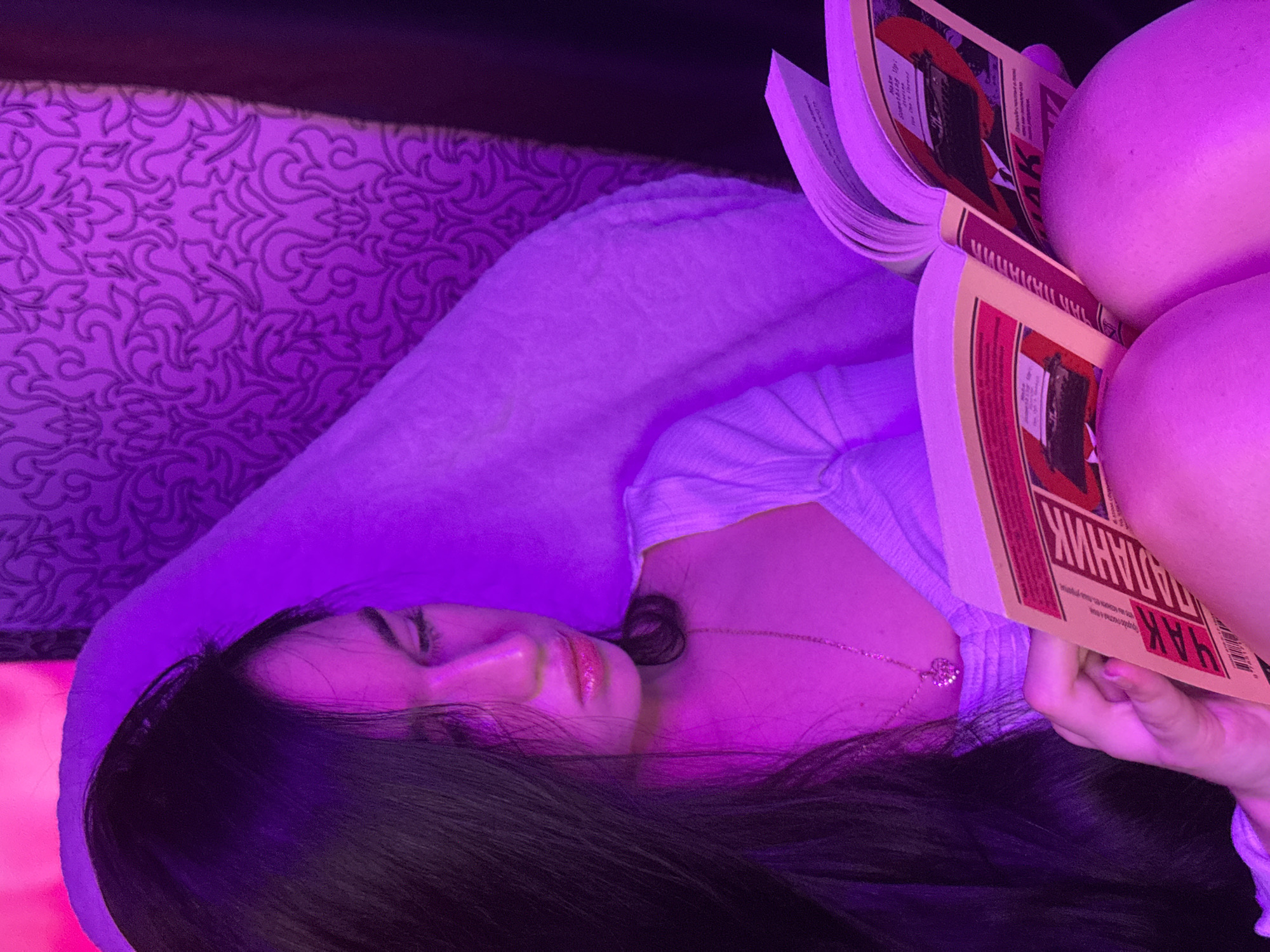 A young woman with long, dark hair is illuminated by purple light as she reads a book. She is wearing a white, ribbed top and a delicate necklace with a heart-shaped pendant. The book is open, and the cover features red and black graphics, with Cyrillic text visible. She is holding the book with her left hand, and her right leg is bent, supporting the book. The background consists of a patterned wallpaper and a dark curtain. The scene appears to be indoors, possibly in a residential setting. The provided image lacks sufficient visual cues to determine a specific location within Novosibirsk, Russia.