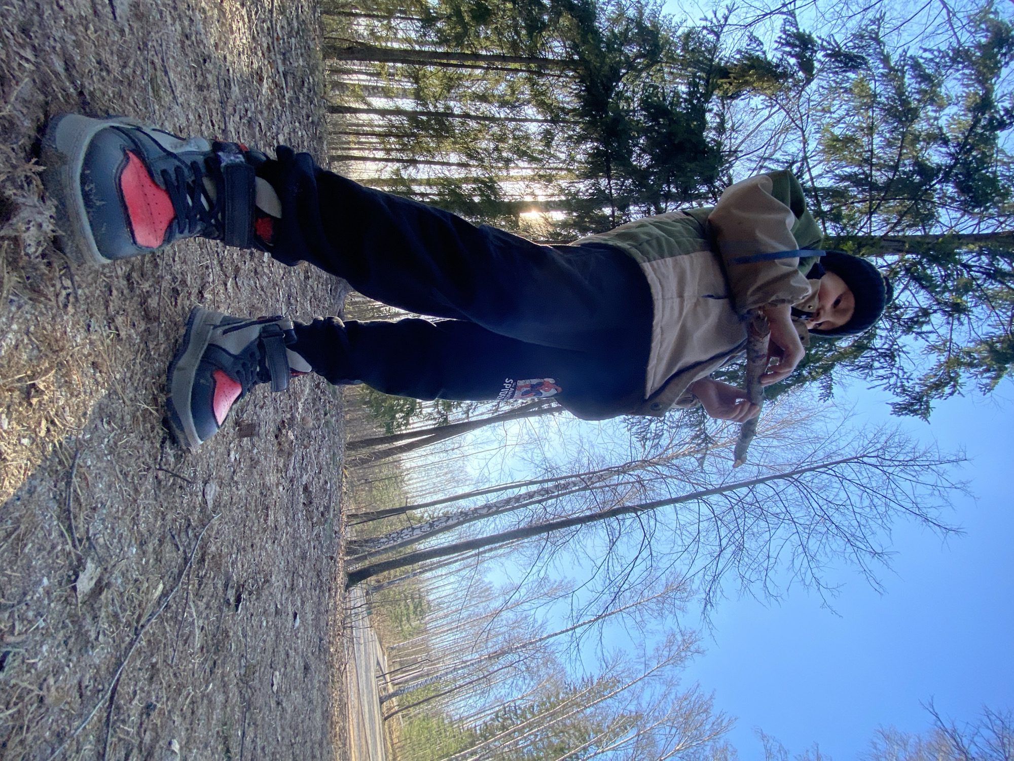 A person is standing in a forest, possibly in Noviy, Russia. The person is wearing a jacket, black pants with a red emblem on the leg, red and grey shoes, and a black hat. They are holding a stick. The ground is covered in leaves and twigs. Trees surround the person with a clear blue sky visible overhead.
