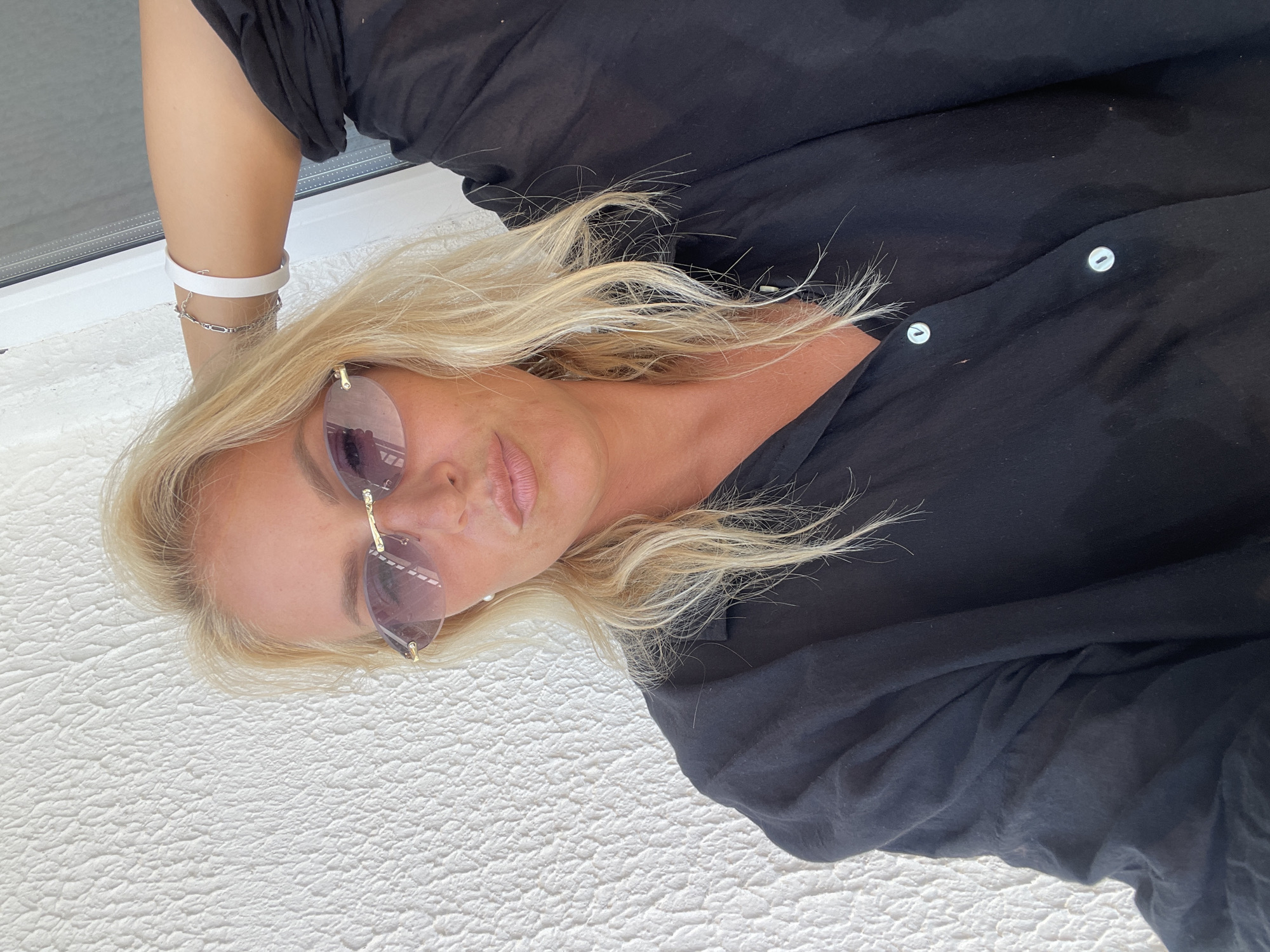 A blonde woman with sunglasses and a black shirt poses for a selfie against a textured white wall. The woman is wearing light-colored lipstick and has her left arm raised behind her head. A silver bracelet and a white band are visible on her left wrist. The background wall has a rough, stucco-like texture. The image appears to be taken outdoors, possibly on a balcony or patio, given the bright lighting and the edge of a window frame visible on the right side. No specific landmarks or urban features are identifiable to place it within Giachrypsh, Abkhazia.