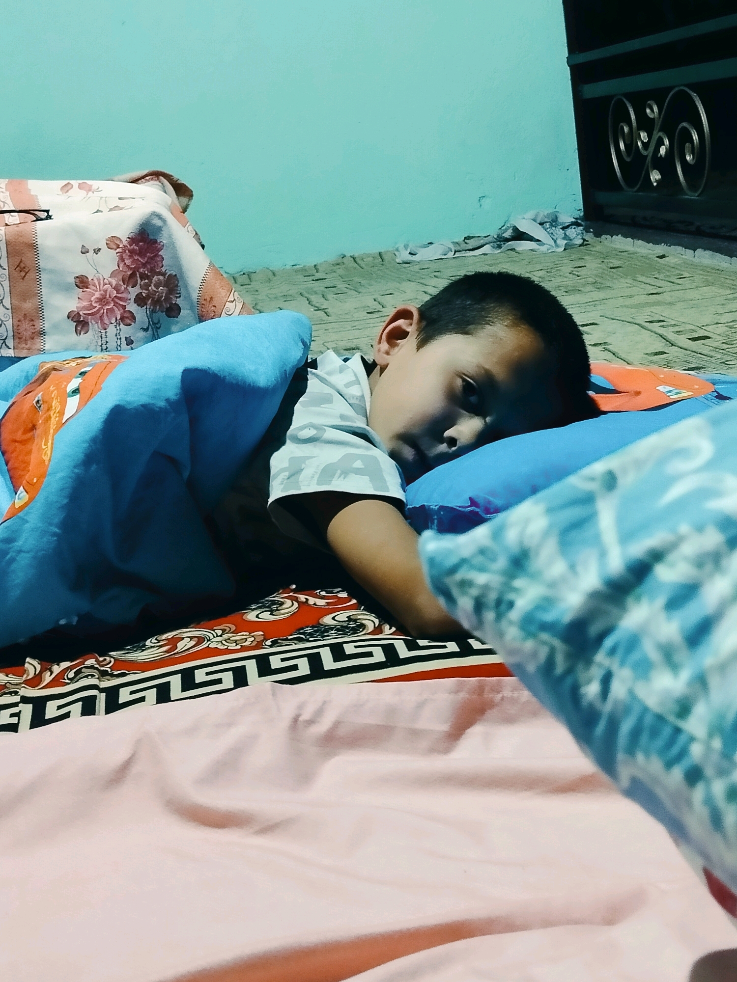 A young boy is lying on his side, asleep on a bed. He is covered by a blue blanket and has a pillow under his head. The bed is covered with a patterned sheet and a pink blanket. The room appears to be lit by artificial light. The background shows a light blue wall and a window with a decorative metal grill. Based on the typical architecture and interior design, this scene is likely located in a residential area of the city of Көлкент, Kazakhstan.