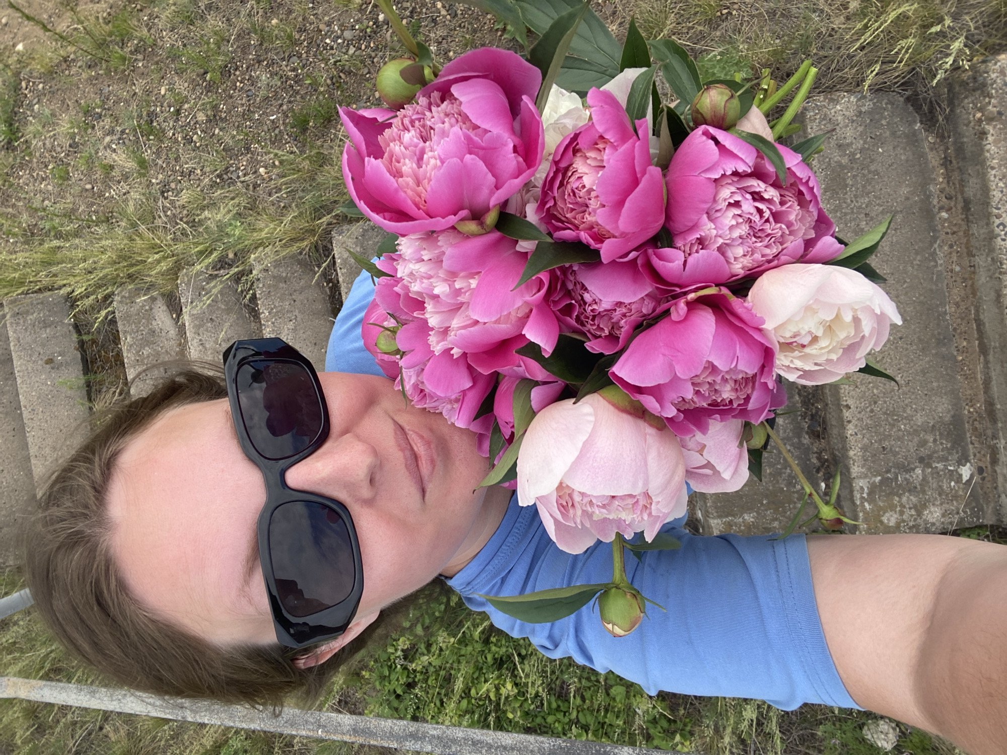 A person wearing sunglasses and a blue shirt holds a bouquet of pink and white peonies. They are standing on a concrete stairway next to a grassy embankment. The scene appears to be outdoors in a natural setting.