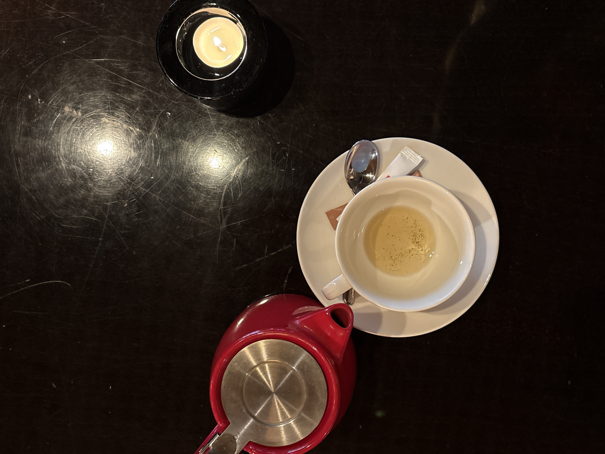 An overhead shot depicts a dark, possibly wooden or laminate surface on which several objects are arranged. A small, black candle holder with a lit candle sits in the upper-left quadrant. Near the center, a white saucer holds a white teacup filled with liquid. To the right of the cup is a silver spoon and a small white packet. In the bottom-left quadrant, a red teapot is positioned. The scene is lit by the candle and a light source above, casting shadows. There are no people visible. There is no explicit indicator the image was taken in London, United Kingdom.