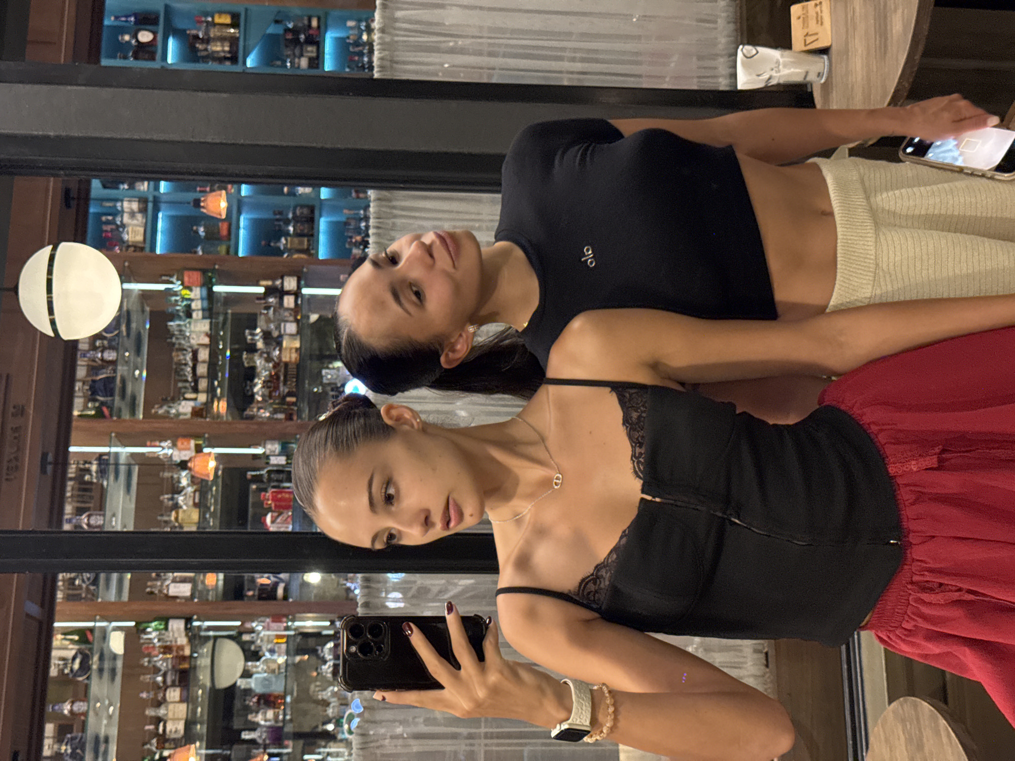 Two women are posing for a selfie in Phuket City Municipality, Thailand, within a bar or restaurant setting. One woman, holding a smartphone, is wearing a black lace-trimmed camisole with a zipper down the front, a silver necklace, a white smartwatch, and a beaded bracelet. She is wearing a red ruffled skirt. The second woman is behind her, wearing a black crop top and beige knitted pants. In the background is a bar with multiple shelves stocked with liquor bottles and various decorative items. A spherical light fixture and a wooden table are also visible.