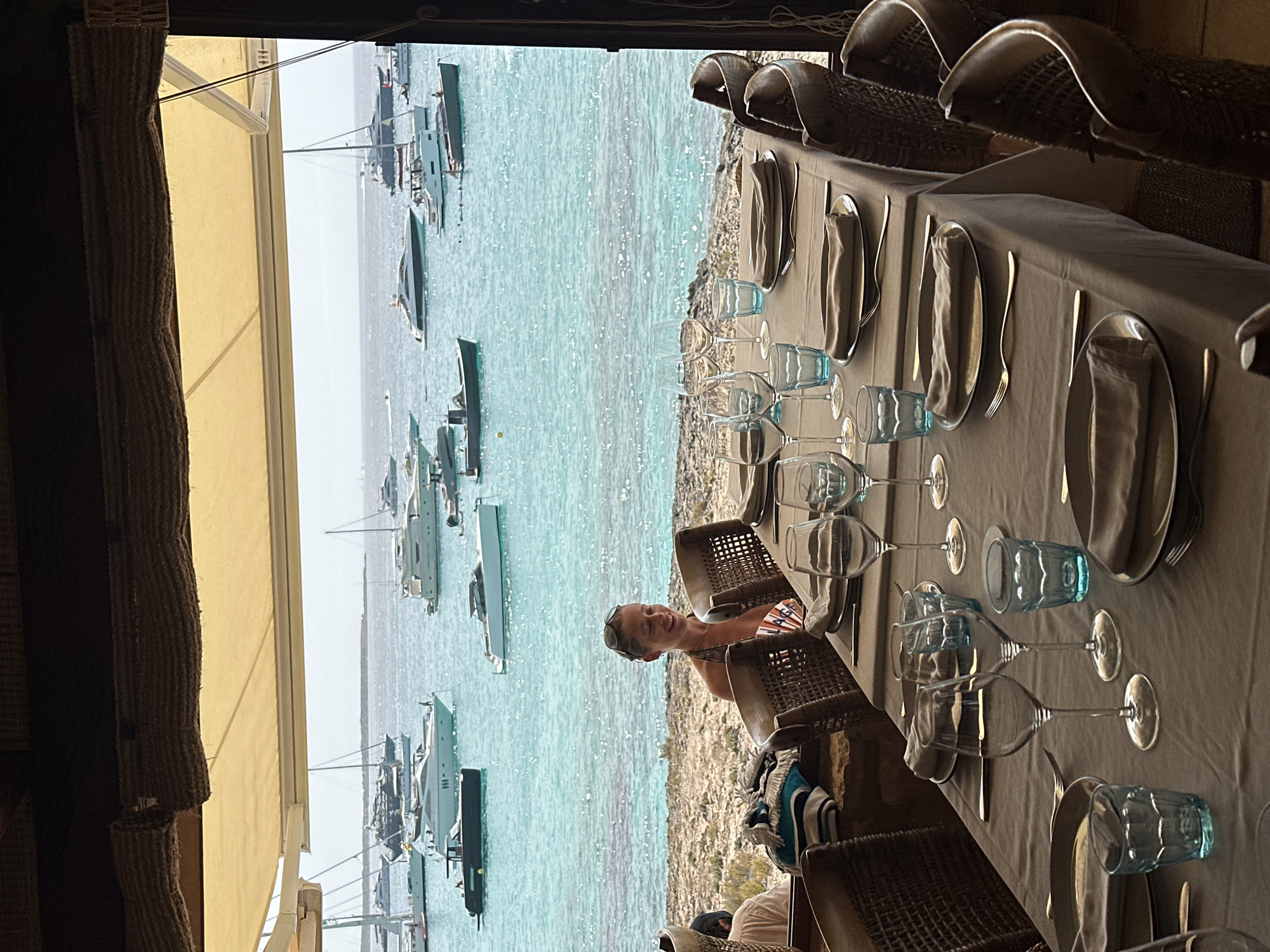 The image depicts an outdoor dining area with a table set for a meal, overlooking a body of water filled with numerous boats. A woman is seated at the table, looking to her left and smiling. The table is covered with a light-colored tablecloth and set with plates, napkins, silverware, wine glasses, and blue tumblers. The chairs surrounding the table appear to be made of rattan. In the background, the turquoise water is dotted with various yachts and smaller boats. The scene is framed by a thatched awning overhead. The environment suggests a seaside restaurant or a similar establishment located by the water.