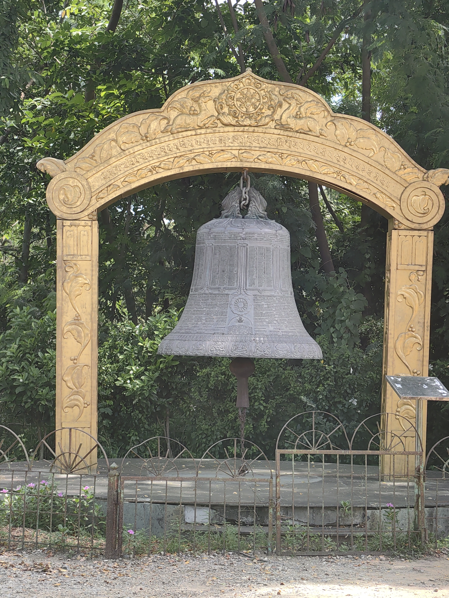 The image shows a large, ornate bell hanging from a golden archway in Lumbini Bazaar, Nepal. The archway is decorated with intricate carvings and religious symbols. The bell itself is made of a metallic material and is covered in inscriptions. In front of the bell is a rusty metal fence, behind which are some small pink flowers and lush green foliage. The background consists of trees and dense vegetation, indicating an outdoor setting. The ground in the foreground is covered with gravel. There are no people visible in the image. The overall scene suggests a spiritual or cultural site.
