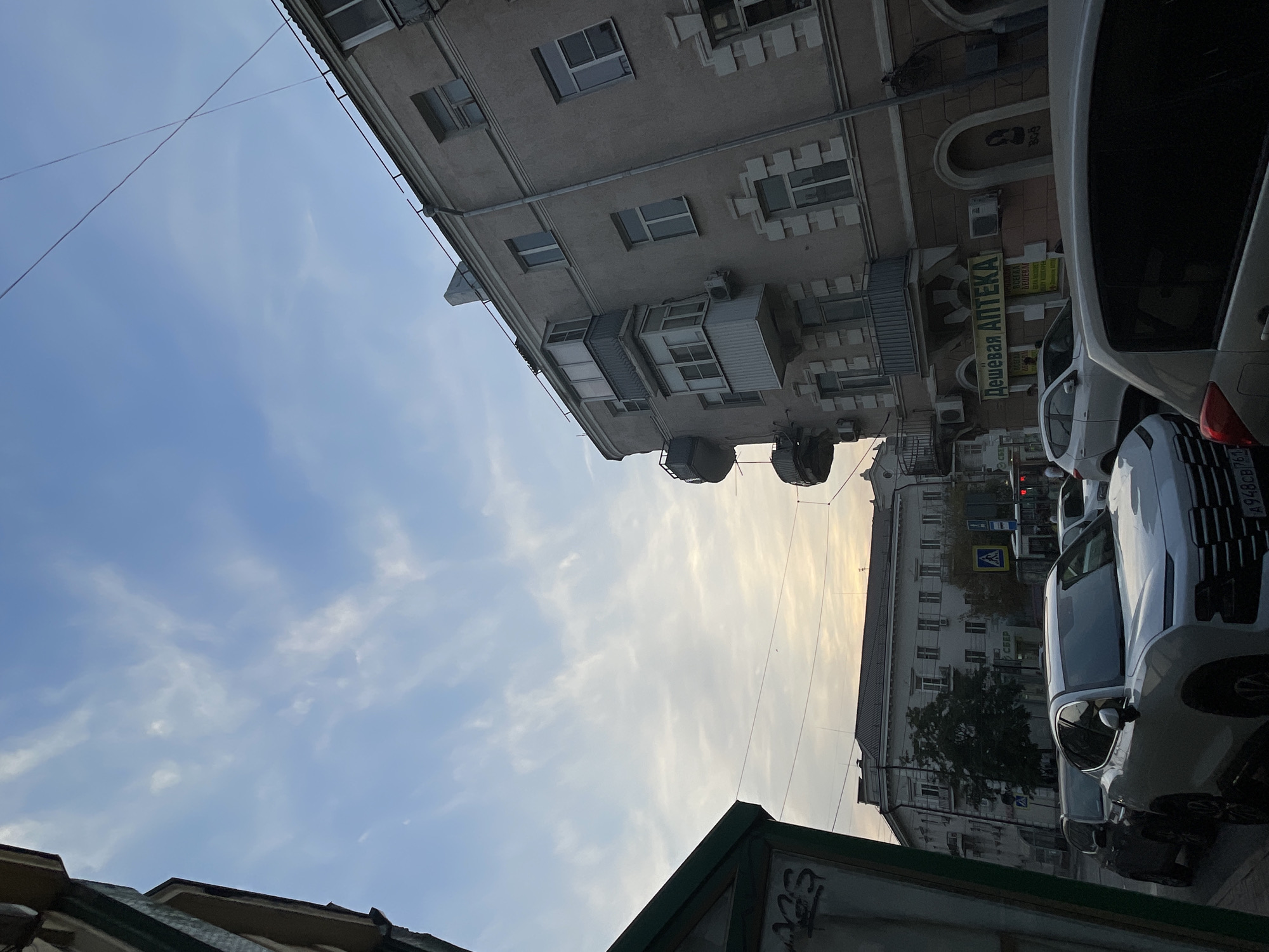 A street scene in Rostov-on-Don, Russia, shows buildings, cars, and a cloudy sky. A light-colored, modern SUV is in the foreground, facing left. To its left, a dark-colored SUV is parked. Behind these, a white hatchback is visible. Further down the street, a light-colored sedan is parked.
The buildings are multi-story, with balconies and windows. A sign in yellow and green reads "Дешевая АПТЕКА" (Cheaper Pharmacy). A traffic light is visible, as are street signs and utility poles with wires. The sky is partly cloudy with a mix of blue and white. The scene appears to be in an urban area.