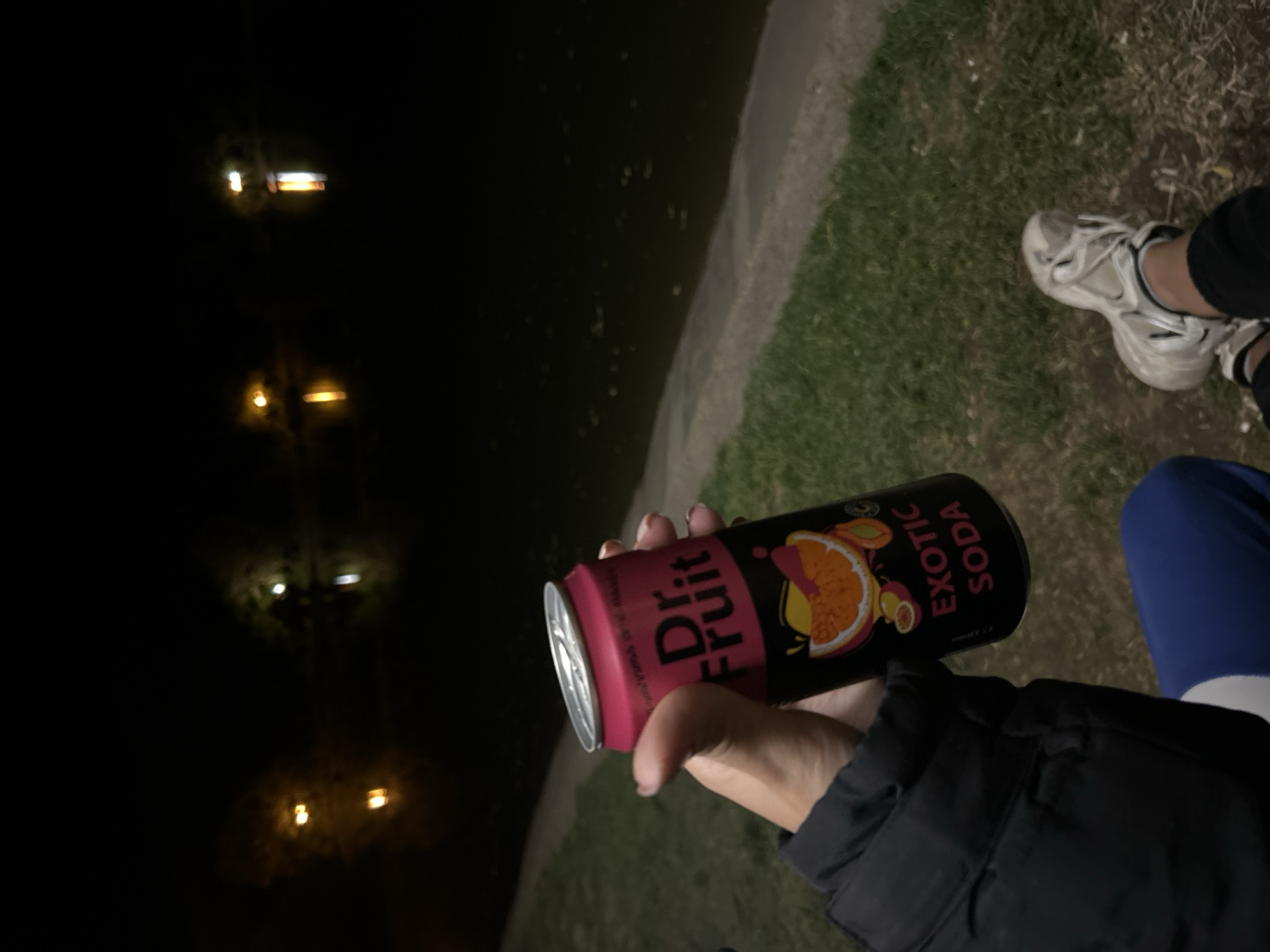 A person is holding a can of "Dr. Fruit Exotic Soda" in their hand. The background shows a dark area, possibly water reflecting lights, and some grass. The person is wearing a dark jacket. White sneakers are visible. The location appears to be outside at night.