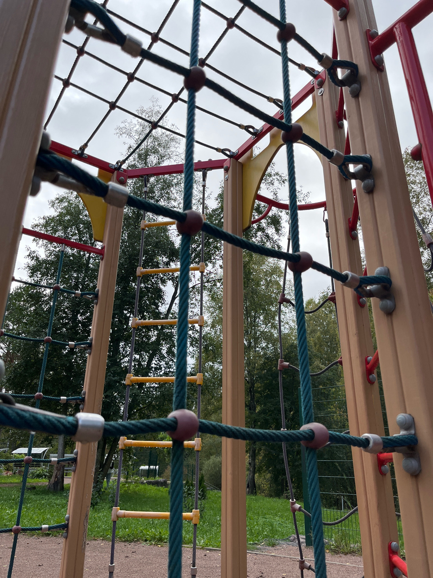 The image shows a close-up view of a section of a playground structure, primarily focusing on the climbing net and wooden support beams. The climbing net is made of thick green ropes interwoven to form a grid. Red, brown, and silver spherical connectors are visible where the ropes intersect and attach to the structure. The support beams are made of light brown wood with some red accents. In the background, a ladder with yellow rungs is visible through the net, and beyond that, lush green trees and a cloudy sky can be seen. The ground appears to be a mix of gravel and grass. The scene is set in an outdoor park.