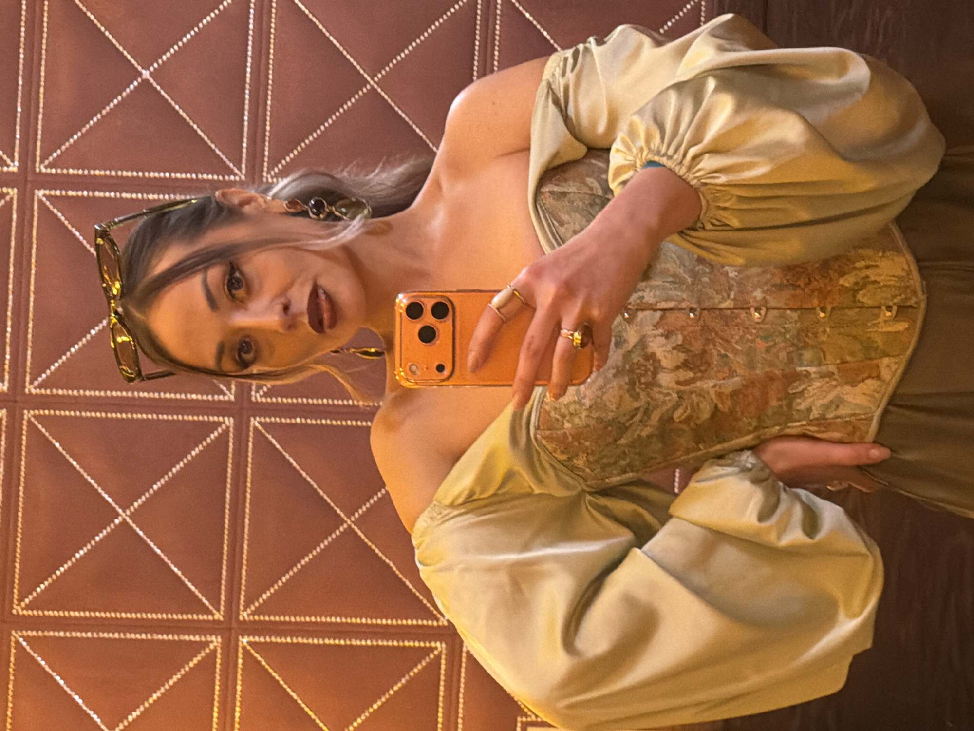 A woman is taking a selfie in front of a brown, diamond-patterned wall. She is wearing an off-the-shoulder sage green dress with puffy sleeves and a floral corset. Her blonde hair is styled with sunglasses perched on top of her head. She is holding an orange smartphone in her right hand, displaying it to the camera. Several rings are visible on her fingers. The background is out of focus. I have no information about a location in Krasnoyarsk, Russia.