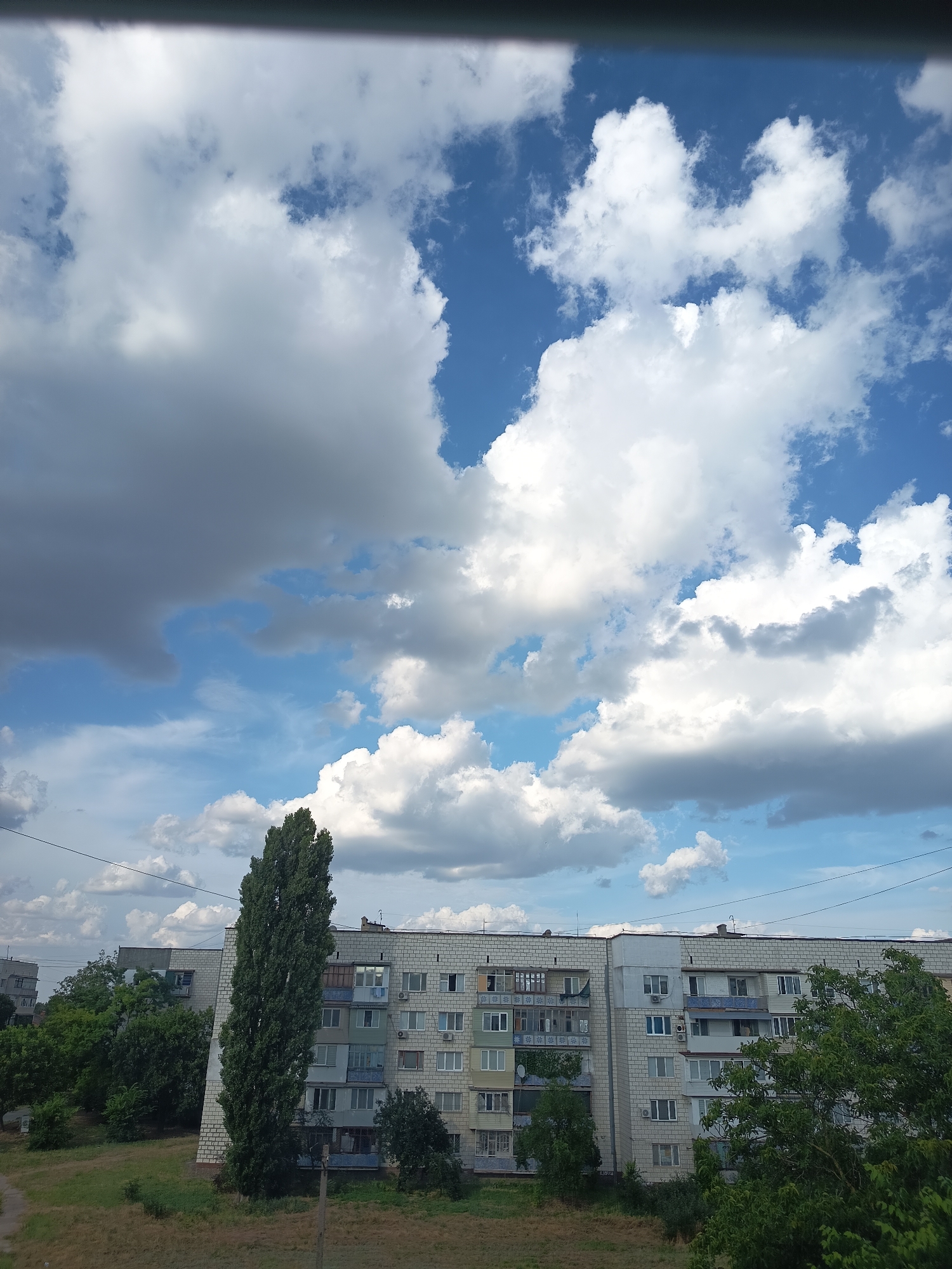 The image captures a view of a multi-story apartment building with a light-colored brick facade and numerous windows and balconies. A tall, slender tree stands prominently in front of the building. The sky is filled with a mix of white and gray clouds against a blue backdrop. In the foreground, a patch of dry grass and some green trees are visible. The scene appears to be an urban environment in Izmail, Ukraine, likely taken from an elevated position, possibly a window or another building. There are no people or specific actions depicted. The overall atmosphere is overcast with a bright sky.