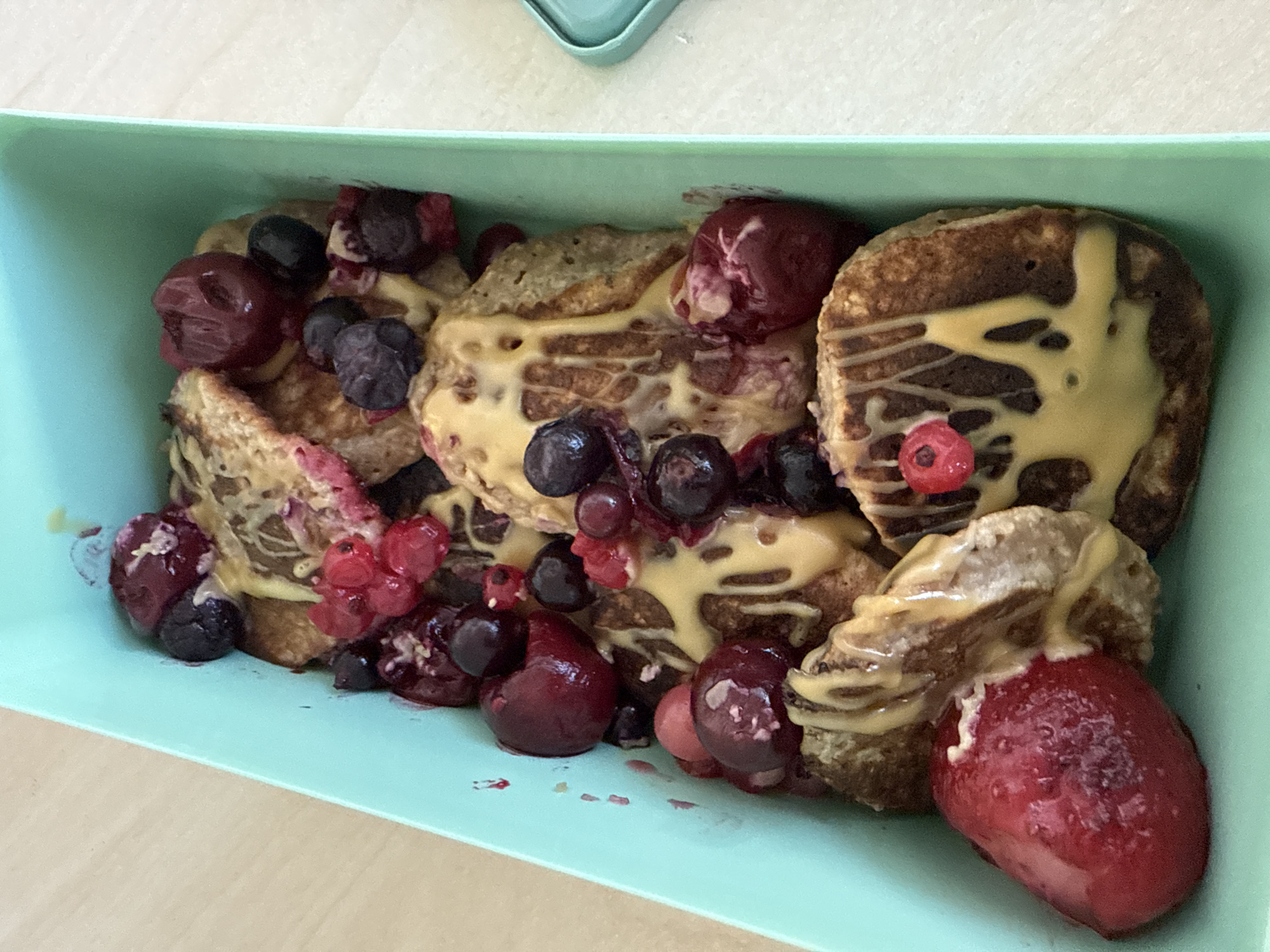 The image shows a teal-colored container filled with pancakes topped with a light-colored drizzle, blueberries, and cherries. The container is sitting on a light brown surface. There is no indication in the image of Sofia, Bulgaria. There are no people in the picture.
