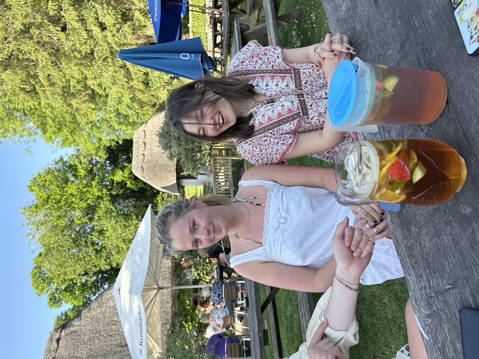 Two women are seated at a wooden picnic table outside. One woman is wearing a white dress and looking at the camera, and the other is wearing a floral patterned dress and smiling with her eyes closed. They are holding hands. There are two pitchers of tea with fruit slices on the table. In the background, there is a thatched-roof building, people seated at tables under umbrellas, and green trees. The location is Unknown, Unknown.