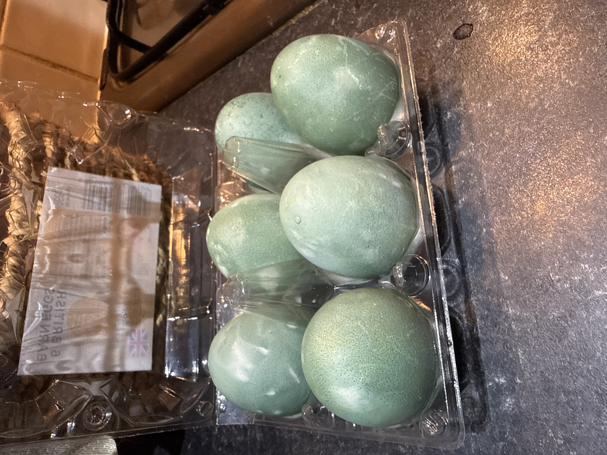 A close-up photograph captures a half-dozen pale greenish-blue eggs nestled in a clear plastic egg carton. The carton rests on a dark gray countertop. Partial view of kitchen counter with a gas stove above and to the left of the plastic tray of eggs. A small Union Jack is visible on the package insert. No people are visible. The location in Great Yarmouth, United Kingdom is inferred from the packaging.