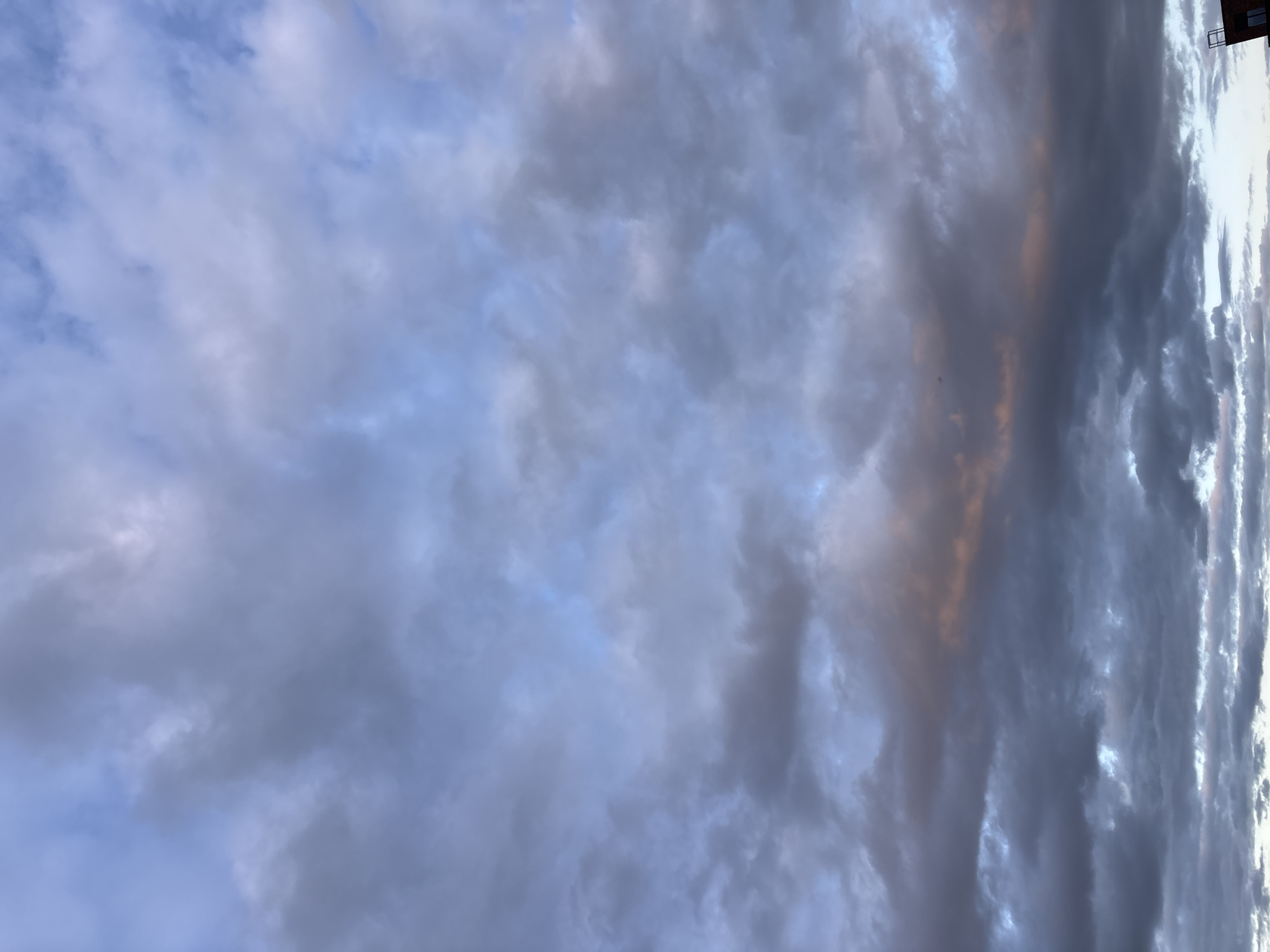 A wide-angle, low-angle shot depicts a sky filled with predominantly gray, cumulus clouds, with hints of pale blue visible in the thinner sections. A subtle orange glow emanates from the horizon line, suggesting either sunrise or sunset. In the lower right corner of the image, the top of a brick building with a dark, angular structure on its roof is visible, indicating an urban or suburban environment. The scene appears to be captured in Zvenigorod, Russia, at an atmospheric dusk or dawn. There are no people, animals, or distinct actions captured in the frame.
