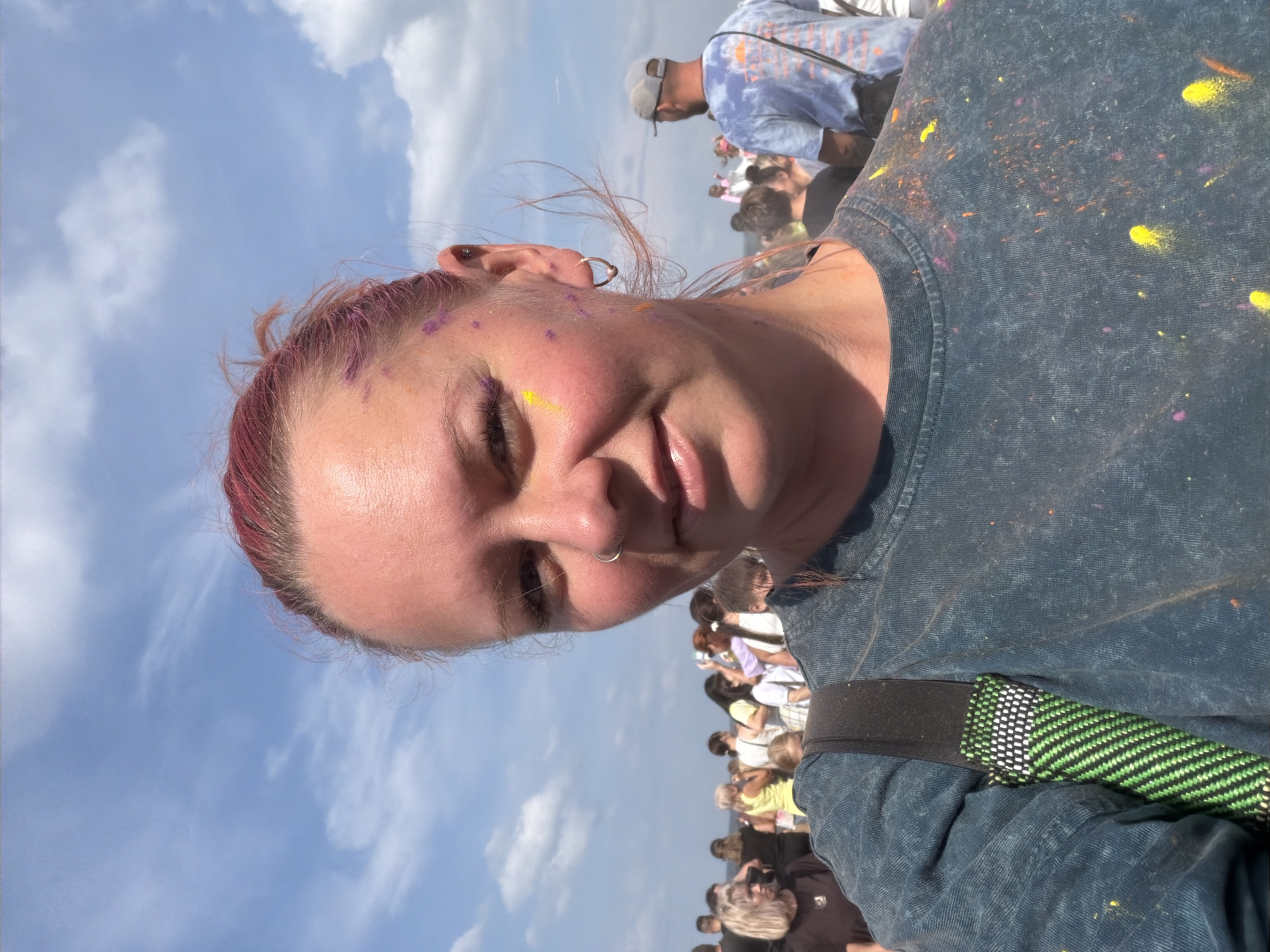 The image depicts a woman with pink hair, her face partially covered in colorful powder, likely from a festival or event. She is wearing a dark, possibly denim, t-shirt speckled with various colors of powder, including yellow, purple, and orange. A green and black striped strap of a bag is visible over her left shoulder. Her left eyebrow appears to have a streak of yellow powder. She has a silver nose ring and a small hoop earring in her left ear.
Behind her, a crowd of people are visible, suggesting an outdoor gathering. The sky is blue with scattered white clouds. The general atmosphere seems festive and energetic. The colors on the woman and her clothing are indicative of a Holi festival or a similar color-throwing event.
Although the specific location in the city of Chaikovsky, Russia is not explicitly identifiable from the visual cues alone, the context of a large outdoor gathering with colorful powder suggests a public event or festival. Without further information or landmarks, a precise location within the city cannot be determined.