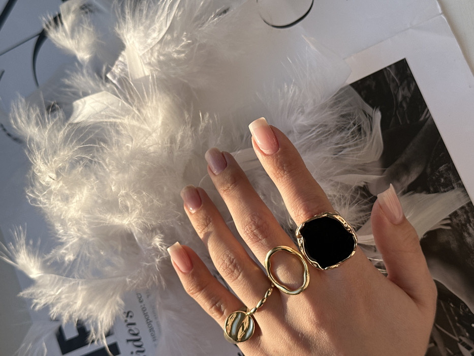 The scene depicts a close-up view of a human hand adorned with three gold-toned rings, resting upon a surface covered with white feathers and printed material. The hand is positioned with its fingers extended, revealing the dorsal side, and has a light-to-medium skin tone. The fingernails are well-maintained, featuring a light pink base with translucent white tips, consistent with a natural or French manicure style.
On the index finger, a gold-toned ring features a circular, slightly convex element with a light, possibly iridescent, patterned surface. On the middle finger, an abstract gold-toned ring forms an irregular open oval. On the ring finger, a larger gold-toned ring prominently displays an irregular, organically shaped black stone or enamel inlay, bordered by an unevenly textured gold-toned setting.
The background consists of a dense layer of loose, soft white feathers. Beneath the feathers, sections of a printed page, likely from a magazine or book, are visible. Partially discernible English text reads "iders" near the top left. Below this, Cyrillic text "недорого: ес" is present. Parts of a black and white image, possibly a photographic print, can be seen on the page underneath the feathers and the hand. The scene is illuminated by warm, directional light, casting subtle shadows and highlighting the textures of the feathers and the hand. This image is situated in the city of Chaikovsky, Russia.