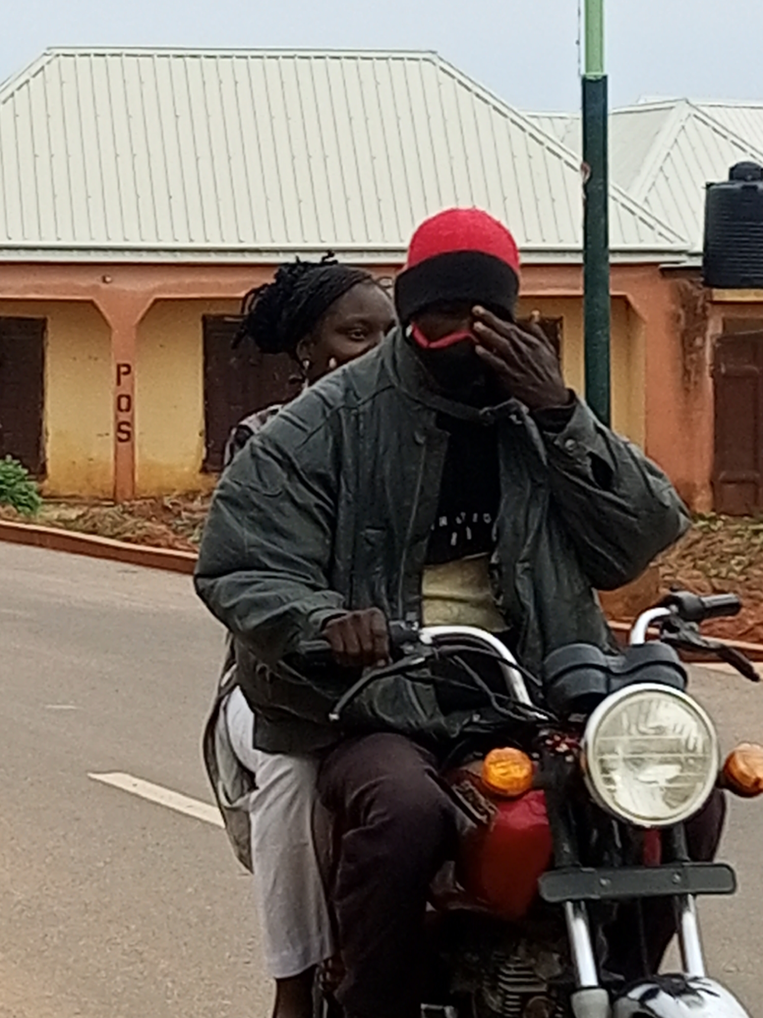 The image depicts two individuals riding a motorcycle on a paved road. The person in the foreground, driving the motorcycle, is wearing a dark jacket, a black shirt, a red cap, and a pink face mask. They are covering their eyes with their right hand. The second individual, a passenger, is seated behind the driver and appears to be wearing white trousers and a dark top. The motorcycle has a red fuel tank and a prominent round headlight.
In the background, a building with a light-colored, corrugated metal roof is visible. The building has an orange facade with dark window openings. A vertical pole, painted green and black, stands to the right of the building. A black water tank is mounted on top of the building on the far right. The phrase "POS" is written vertically on a pillar in front of the building. The overall environment appears to be an urban or semi-urban setting. The scene is captured in daylight, with overcast sky conditions.