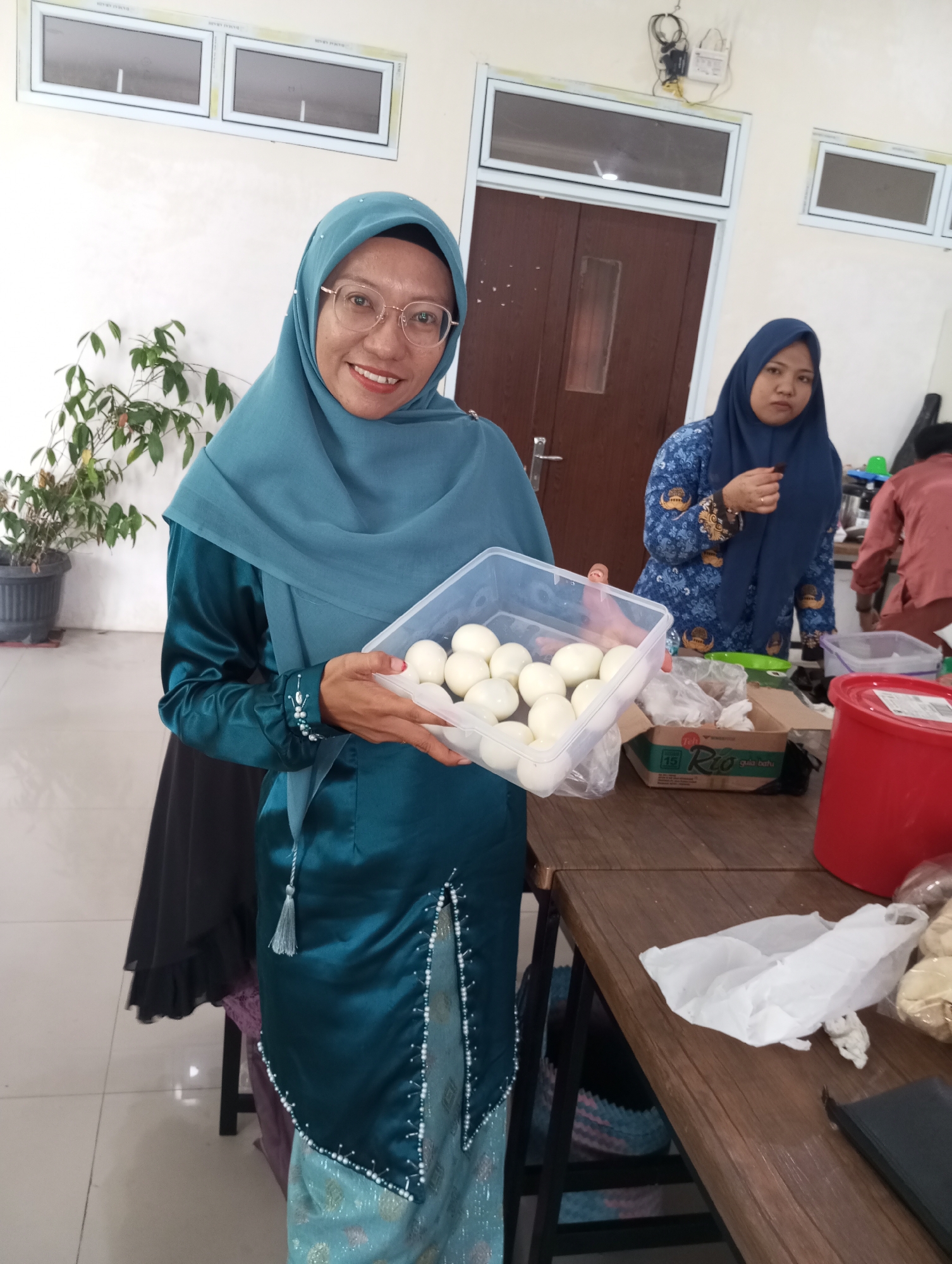 In the city of Pematang Reba, Indonesia, a woman wearing a teal hijab and a matching teal satin dress stands indoors. She holds a clear plastic container filled with numerous hard-boiled eggs. Her dress is adorned with white beadwork along the front slits and cuffs. Behind her, another woman in a blue hijab and a patterned blue top is visible, also in an indoor setting. The environment appears to be a room with light-colored walls, large windows, and a wooden table in the foreground laden with various items including a red bucket, plastic bags, and a cardboard box. A plant in a dark pot is situated to the left of the foreground woman.