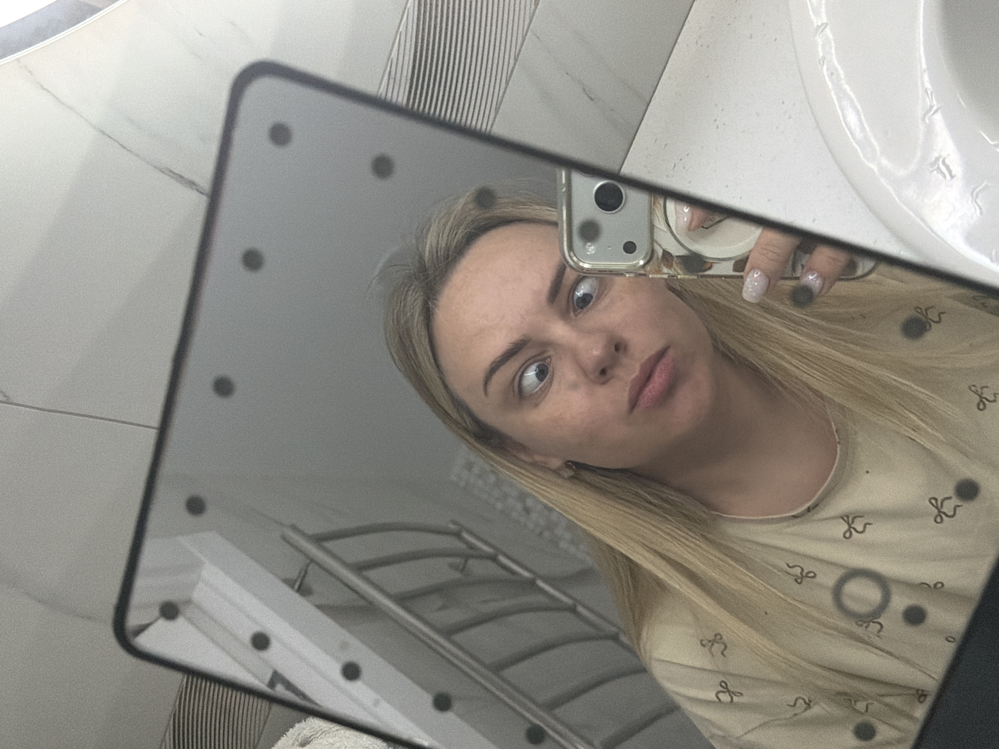 A person with blonde hair is taking a selfie in a mirror, seemingly in a bathroom. The mirror has lights embedded around its edge. The person is holding a smartphone with a case, and the camera is visible. In the background, a bathroom sink, towel rack, and striped tiles are visible. No information about the location in Votkinsk, Russia is apparent in the image.