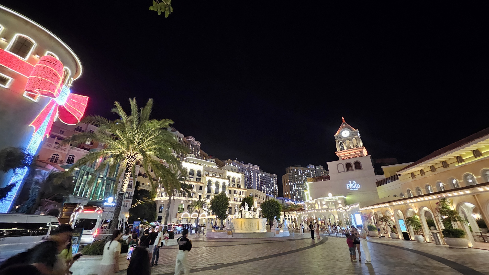 This is a wide-angle, eye-level, nighttime photograph of a plaza in Sun Premier Village, Phú Quốc, Vietnam. The plaza is illuminated by numerous lights, creating a festive atmosphere. In the foreground, a paved walkway curves through the scene, with scattered groups of people walking and standing. On the left side of the image, a large building is decorated with a giant, illuminated red bow, resembling a gift. A palm tree stands prominently in front of this building.
Further into the plaza, a circular fountain with statues is visible, surrounded by more buildings with arched windows and facades. A clock tower with a lit clock face stands on the right side of the scene, and below it, a sign reads "La Festa Studios." The architecture is reminiscent of European styles, with multiple levels and ornate details. In the background, taller apartment or hotel buildings are illuminated. The sky is dark, indicating it is nighttime. The image captures a lively urban environment at night, likely a tourist destination or entertainment complex.