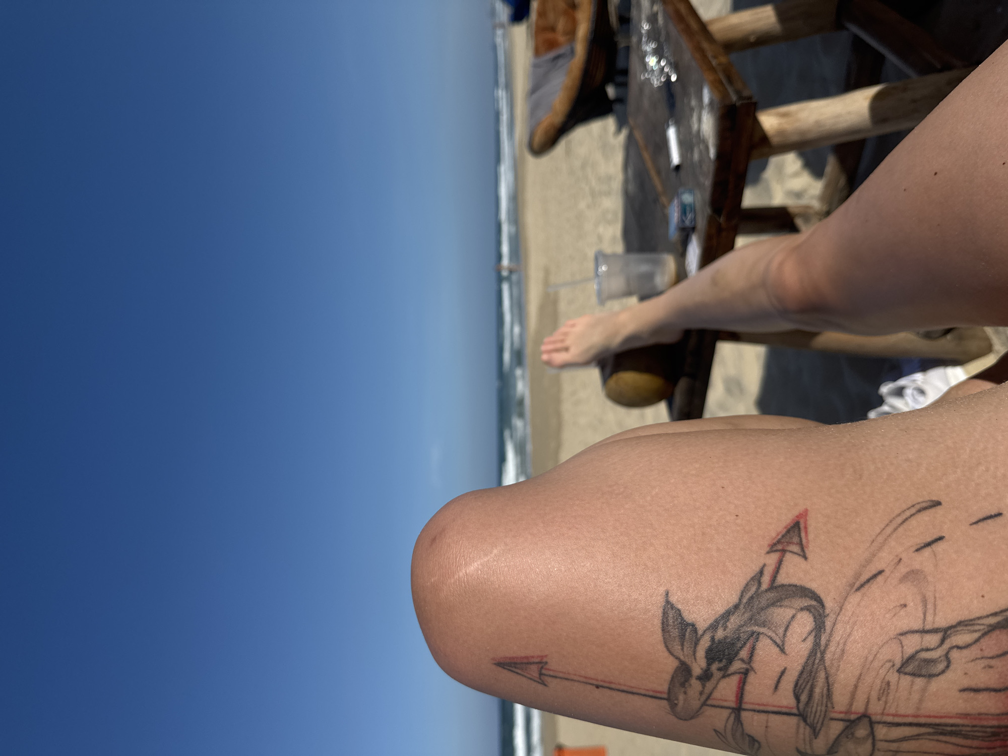 A close-up, angled shot depicts a person's leg with a tattoo of an anchor and a few other objects and scenes in the background. The leg with the tattoo is in the lower right corner of the frame. The tattoo features an anchor with an arrow at the top and a fish-like creature. To the left of the leg is a view of a beach with the sea. A small wooden table with items on top is to the right and slightly behind the leg. In the distance, the blue sky meets the horizon. The location is not explicitly identified as Đà Nẵng, Vietnam in the image.