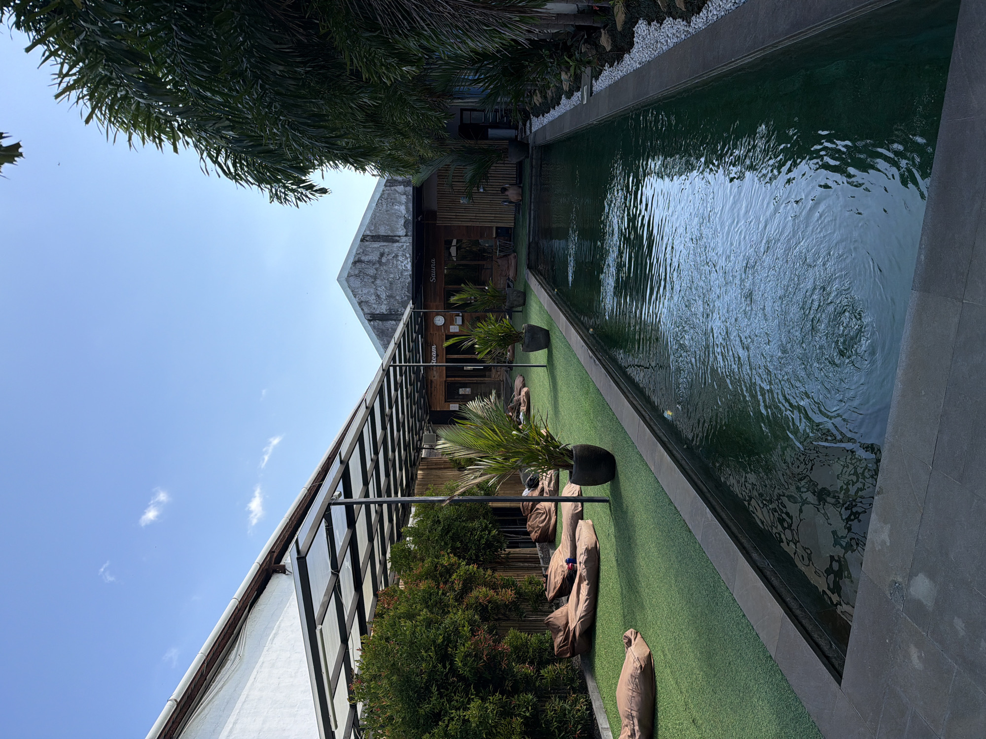 The image depicts an outdoor relaxation area in Denpasar, Indonesia, featuring a long, narrow swimming pool. The pool is surrounded by dark grey paving stones and white pebbles, with lush greenery and palm trees flanking the right side. On the left side of the pool, there is a stretch of artificial grass, dotted with several brown beanbag chairs. Behind the beanbags, a row of dense green bushes lines the area. Further back, a building with a white exterior and a sloped roof is visible, partially covered by a transparent canopy supported by black metal beams. Signs on the building read "Steam" and "Sauna". The sky above is a clear blue with a few scattered clouds. A person is seated on the grass by the pool, facing away from the camera.