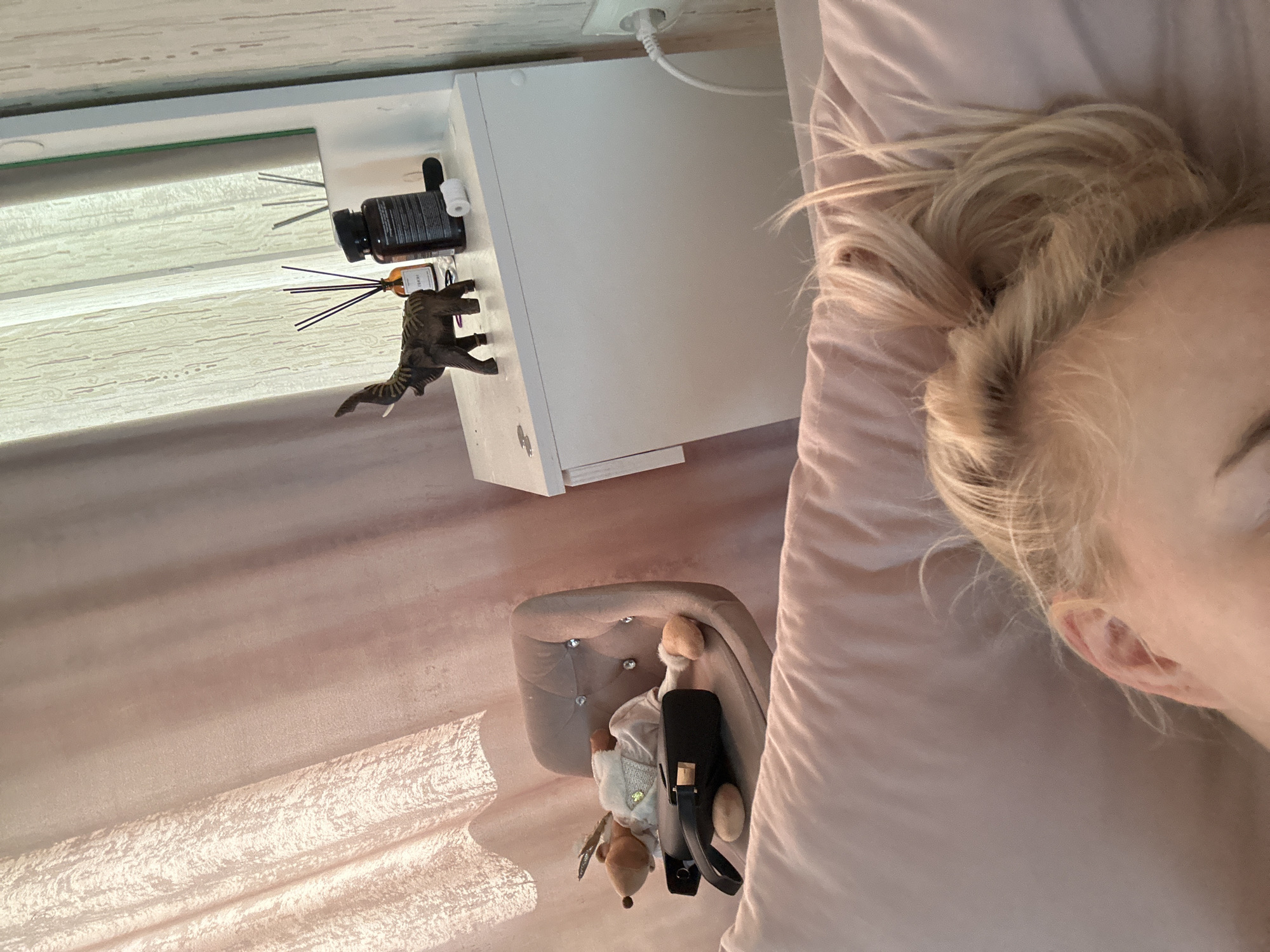 A high-angle shot captures a scene in what appears to be a bedroom. A person with blonde hair is lying on a pink pillow. Adjacent to the pillow, a white nightstand holds various objects, including a black bottle, a diffuser, and a dinosaur figurine. Below the nightstand, a small tufted chair is visible, with a plush animal toy and a black purse resting on it. The room has blinds. There is no specific indicator that this image was taken in Vysokiy, Russia.