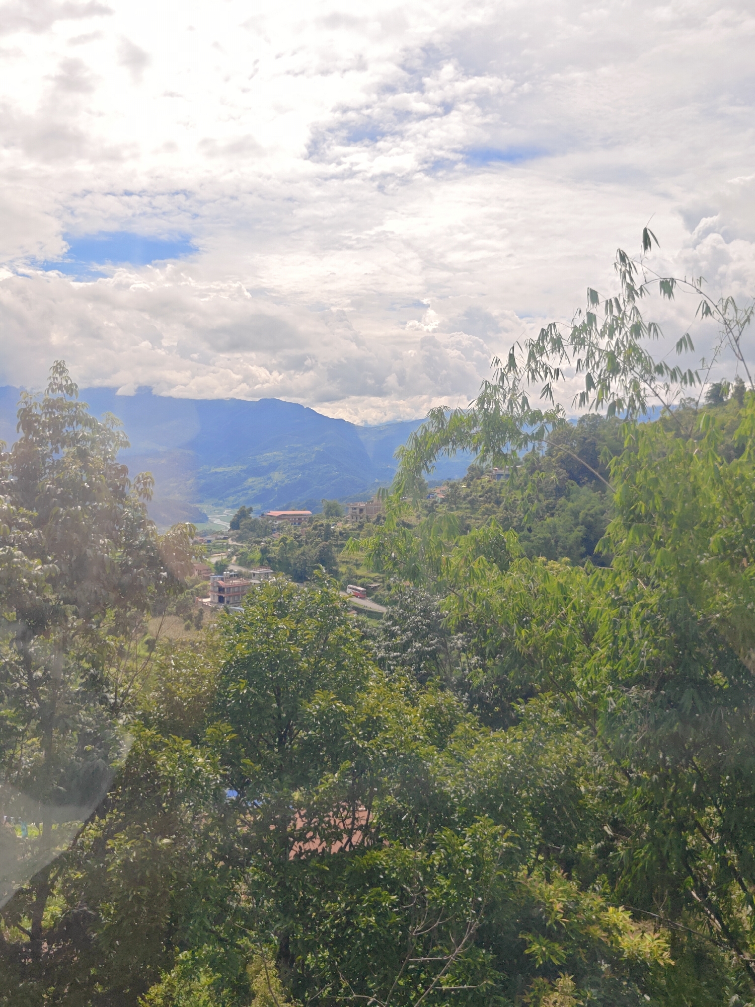 The image shows a scenic view of Pokhara, Nepal, with lush green hills and a valley in the distance. A river winds through the valley, and several buildings, including houses and a larger structure with a red roof, are visible on the hillsides. The sky is partly cloudy, with patches of blue sky visible between the clouds. In the foreground, the branches of several trees, including some with bamboo-like leaves, frame the view. The overall impression is one of a serene and natural landscape, with human habitation integrated into the environment. The scene appears to be a daytime shot, possibly in the late morning or early afternoon, given the lighting conditions. There are no people or animals visible in the image.