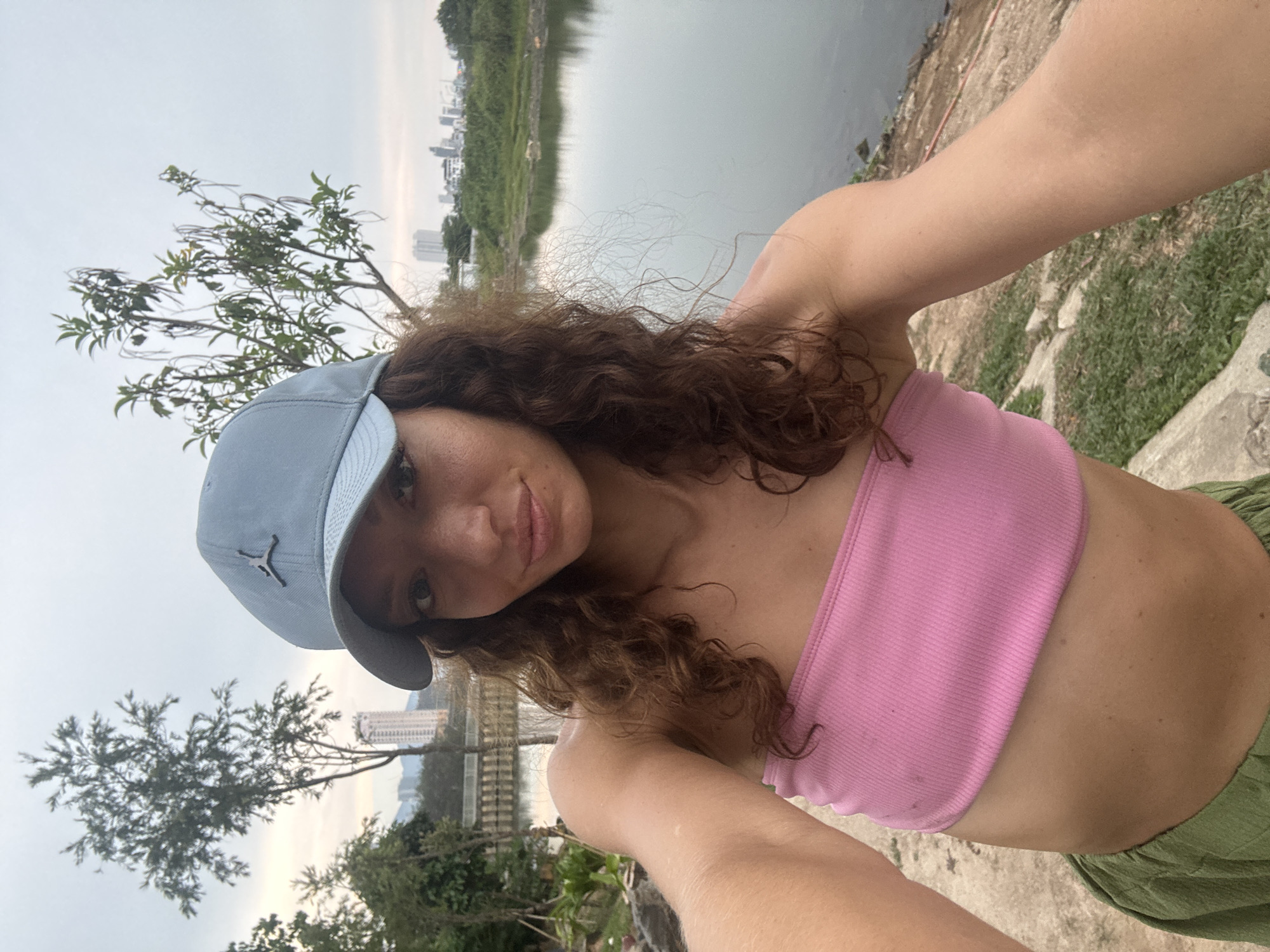 The image depicts a selfie of a young woman wearing a light blue cap with a "Jumpman" logo, a pink tube top, and green pants. She is standing outside, with a body of water and vegetation behind her. In the distance, the skyline of a city is visible. Based on the presence of the body of water and the urban backdrop, it is plausible that the location is somewhere in Đà Nẵng, Vietnam. The woman is holding the camera at an angle, capturing her face, upper body, and the surrounding environment. The sky appears overcast.