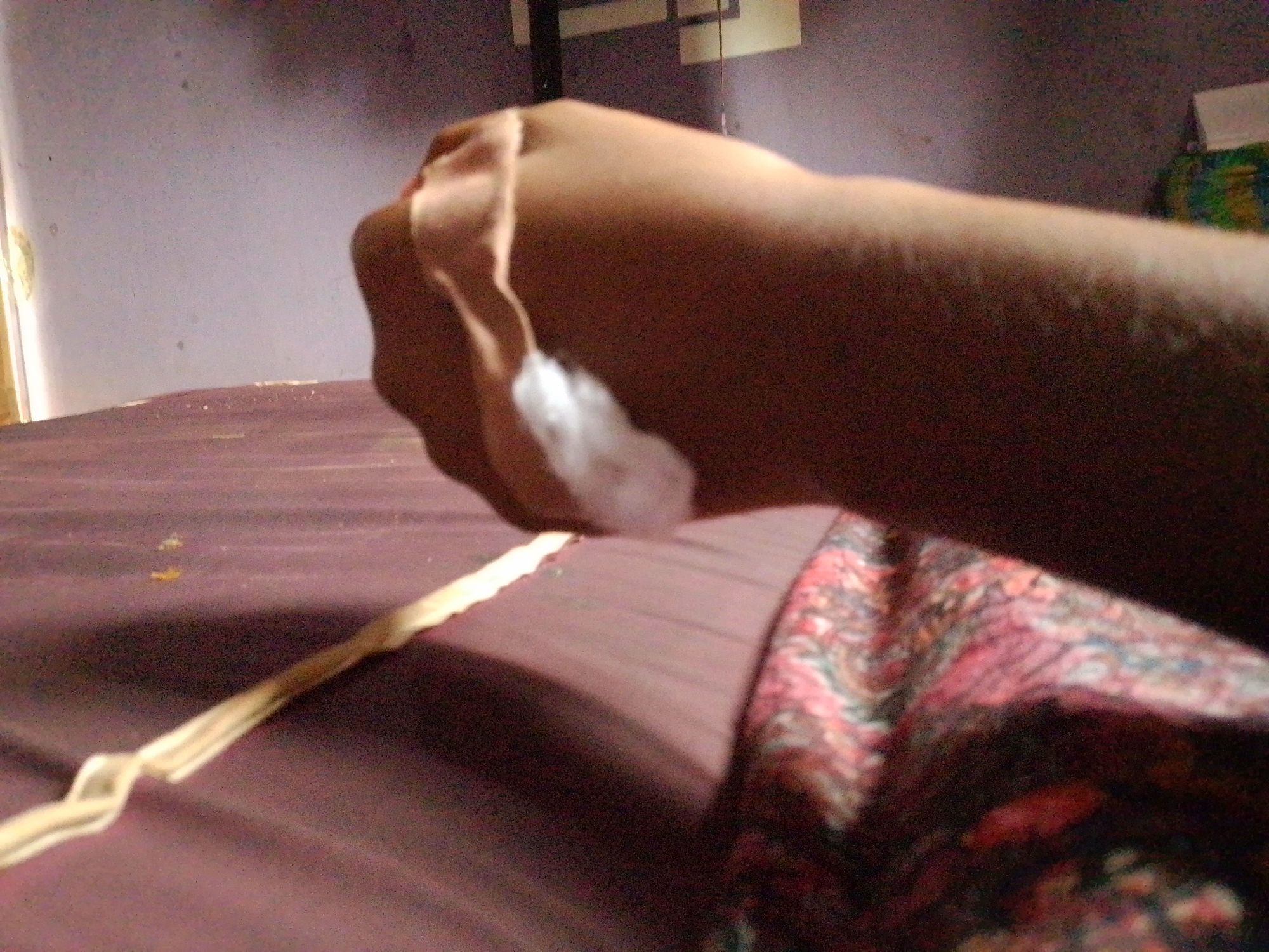The image shows a close-up of a person's arm and hand, with a cotton swab and a piece of string. The arm is extended over a dark-colored surface, possibly a bed, which is covered with a reddish-brown sheet. A patterned fabric, with pink and red floral designs, is visible to the right of the arm. The background is dimly lit and appears to be an indoor setting, with a purple wall and a horizontal element that might be a shelf or frame. There are no discernible elements in the image that would allow for identification of the location within Nsukka, Nigeria. The scene is static and does not depict any ongoing action or interaction.