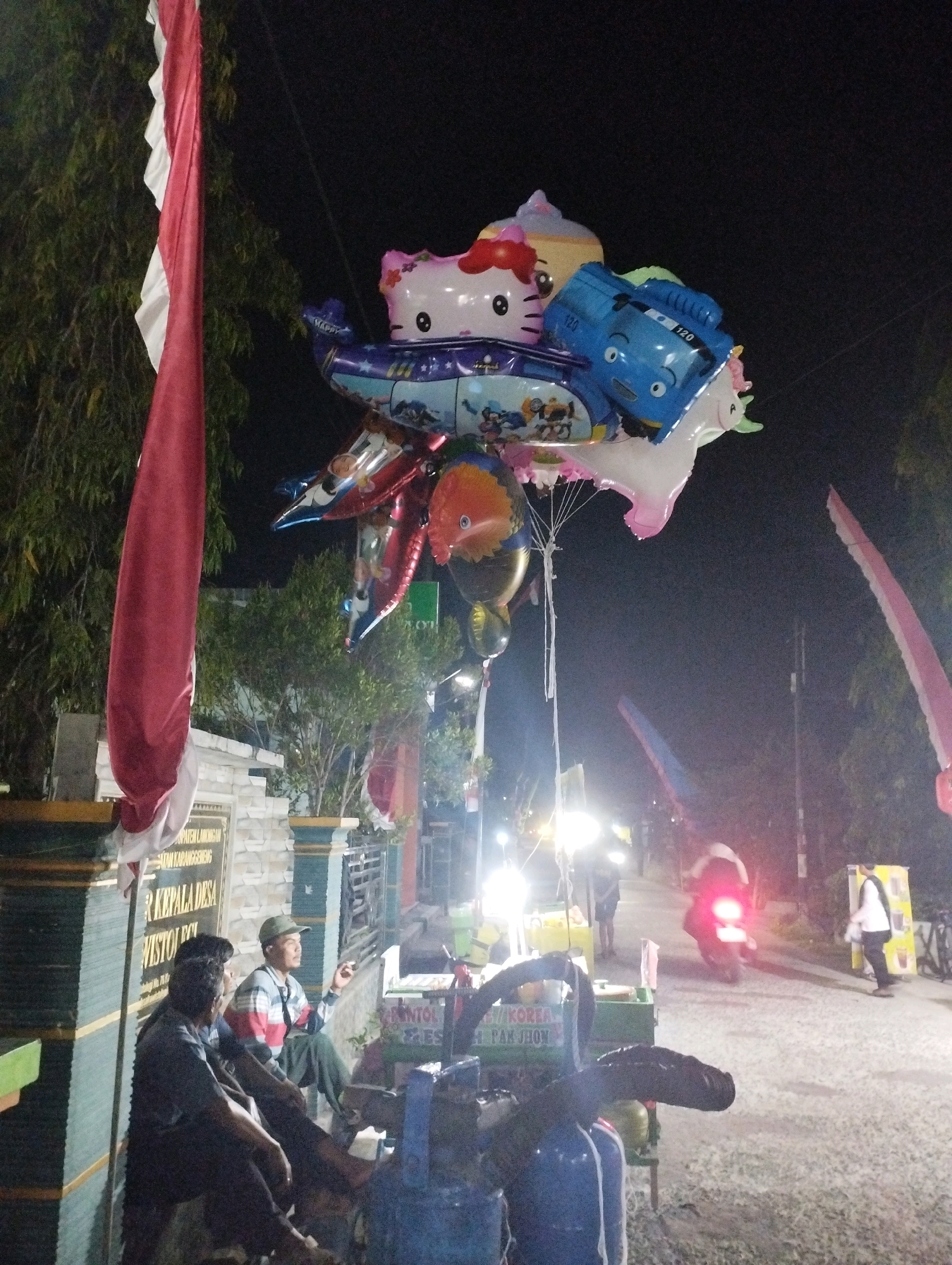 This is a night-time photograph taken in Jagran Kawistolegi, Indonesia, depicting a street scene with several elements. A large red and white flag, likely the Indonesian flag, hangs prominently on the left side of the frame. In the center, a cluster of balloons, including one shaped like Hello Kitty and another resembling a blue bus, are suspended from an umbrella.
Below the balloons, a cart is visible, laden with what appear to be food or drinks. Three men are seated near the cart, two of whom are in the foreground and one further back. The man in the background, wearing a cap, appears to be smoking and looking to the side. A bright light illuminates the scene from the right, suggesting a street lamp or a stall's lighting.
Further down the street, a person is riding a motorcycle, its taillights illuminated. Other individuals are present in the background, some walking and others standing near stalls or structures. The overall environment suggests a bustling night market or a public gathering space. The ground is paved with asphalt, and trees and some buildings are visible in the background. A sign with Indonesian text is partially visible on a wall behind the seated men.