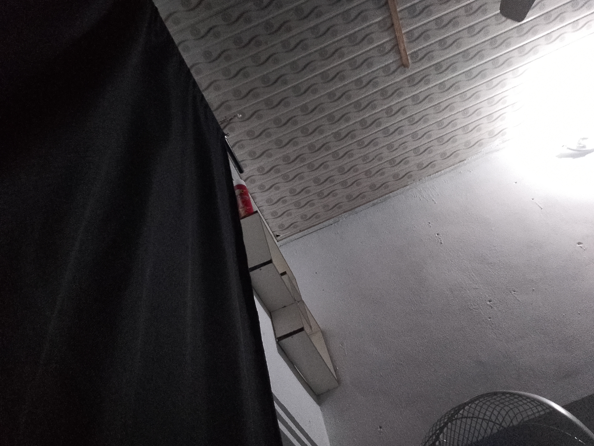 This is a low-angle, wide shot of an indoor space with a textured ceiling and a white wall. A large, dark fabric object, possibly a curtain or clothing, occupies the left side of the frame, obscuring much of the scene. In the upper right quadrant, a bright light source illuminates the ceiling and wall, with a portion of a ceiling fan visible. Attached to the wall, a narrow, box-like structure with a segmented appearance is visible. There are no people or animals present in the image. The scene does not provide any specific indicators of its location within Senti, Nigeria.
