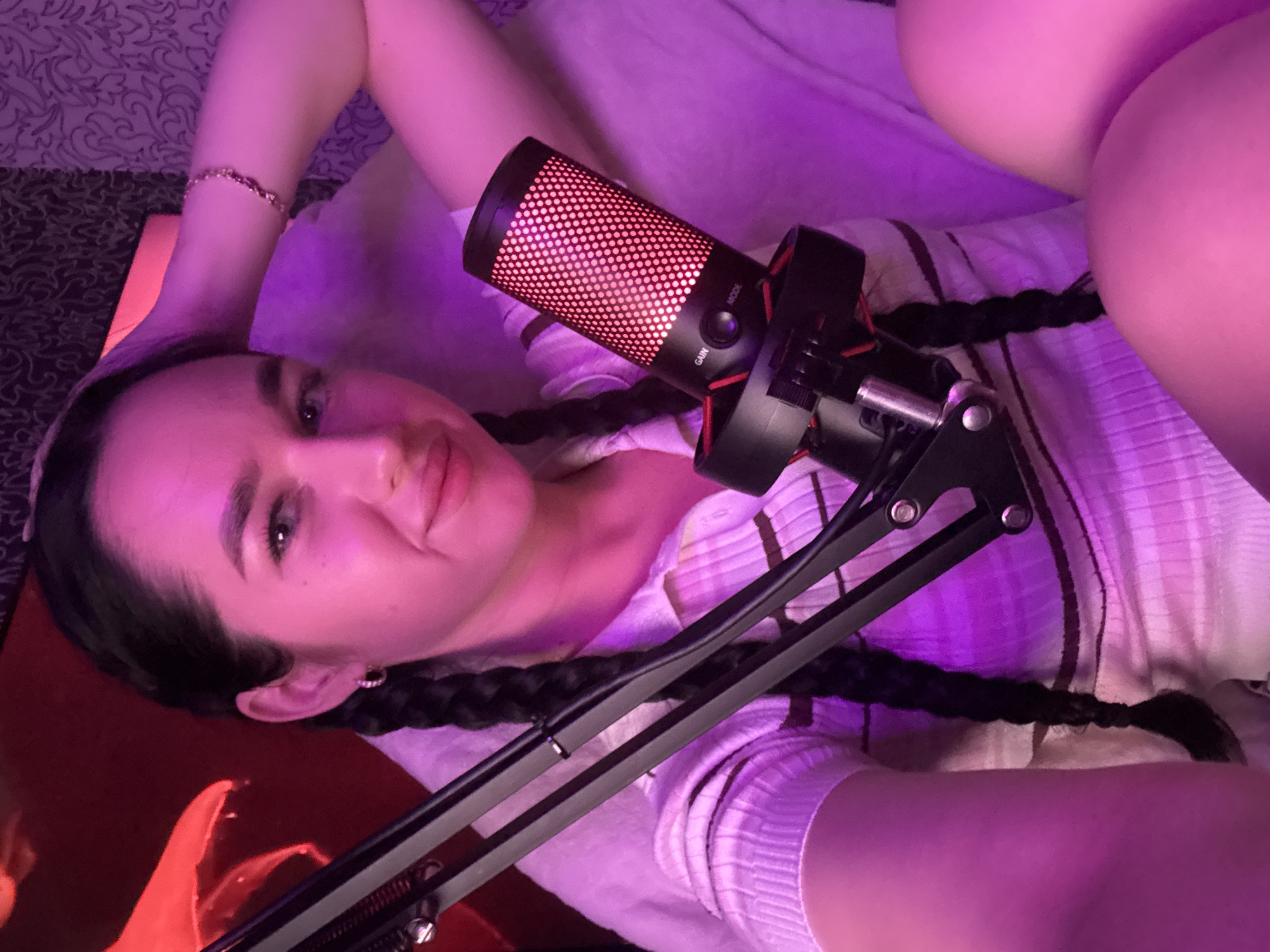 A young woman with dark hair braided into two pigtails is captured in a close-up, selfie-style photograph. She is wearing a pink and white striped collared shirt and has a pink earring in her visible ear. The lighting in the scene is predominantly pink and purple, creating a vibrant and possibly atmospheric setting. A microphone with a red illuminated grille is positioned prominently in front of her. The woman's left hand is raised and resting on the back of her head, and she is wearing a delicate bracelet on her wrist. Her gaze is directed towards the camera with a slight smile. The background appears to be a room with textured wallpaper and possibly a red couch or chair, suggesting an indoor environment. The overall impression is that of a person preparing for or engaging in content creation, such as streaming or recording. The image is devoid of any discernible elements that would specifically place it in Novosibirsk, Russia.