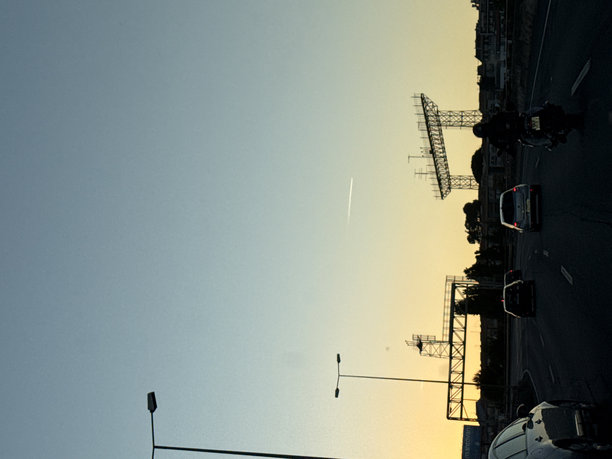 This image captures a scene on a highway in Loures, Portugal, during what appears to be sunset. The sky transitions from a pale blue at the top to a warm yellow and orange hue near the horizon. A distant airplane contrail is visible in the sky. On the left side of the road, a tall street lamp stands prominently.
In the middle ground, a large, industrial-looking metal structure, possibly a billboard support or part of a construction project, dominates the view. Below it, trees and buildings form a silhouette against the colorful sky.
The foreground shows a highway with several vehicles. A silver car is on the left, partially visible. Several other cars are driving on the road, including a dark-colored car in the lane to the right of the silver car, and another car further ahead. On the right side of the highway, a motorcyclist, wearing a helmet, is riding a motorcycle. The road surface is dark asphalt, with visible lane markings. The overall impression is a busy highway scene as day transitions into evening.