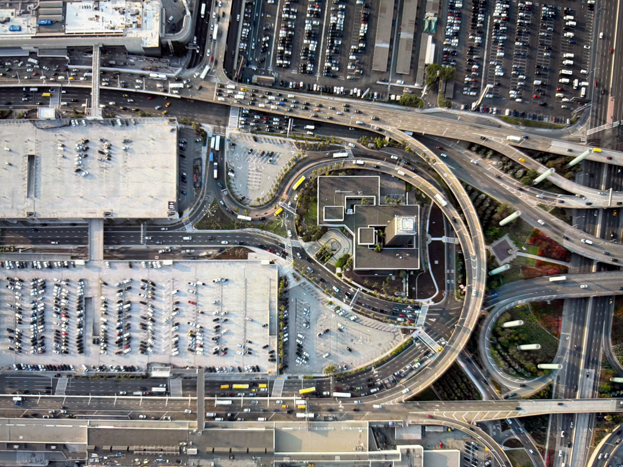 This is an aerial photograph of a large, complex transportation hub, likely an airport or a major interchange, with extensive parking facilities. The scene is dominated by a network of multi-lane highways, overpasses, and on-ramps, with numerous vehicles in motion. Adjacent to the roadways are several large, open-air parking lots filled with a multitude of cars, some in structured rows, others more scattered. There are also large, multi-story buildings that appear to be terminals or parking garages. The overall impression is one of high-volume traffic and a sprawling infrastructure designed to accommodate a significant flow of people and vehicles. The image does not provide any specific visual cues that would allow for the identification of the city of Kyiv, Ukraine.