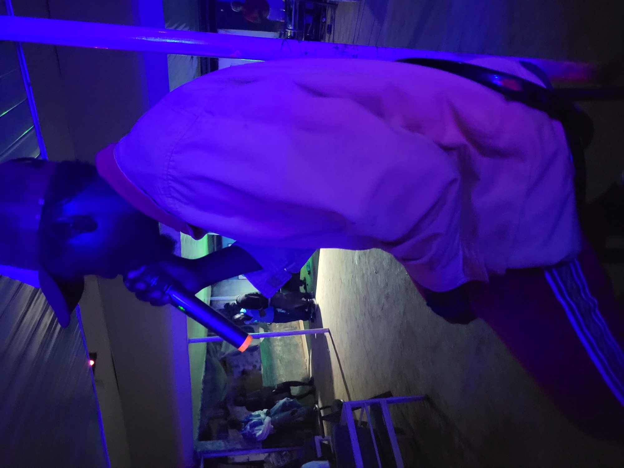 In Agwala, Nigeria, a person wearing a pink shirt and a cap is holding a microphone, seemingly performing or speaking. The scene is illuminated by vibrant purple and blue lighting, casting a dark and moody atmosphere. In the background, several other individuals are visible, some sitting and others standing, suggesting a gathering or event. A white table with some items on it is also present. The overall environment appears to be an indoor space with a rough, possibly unfinished, ceiling. The lighting strongly suggests a performance or party setting.