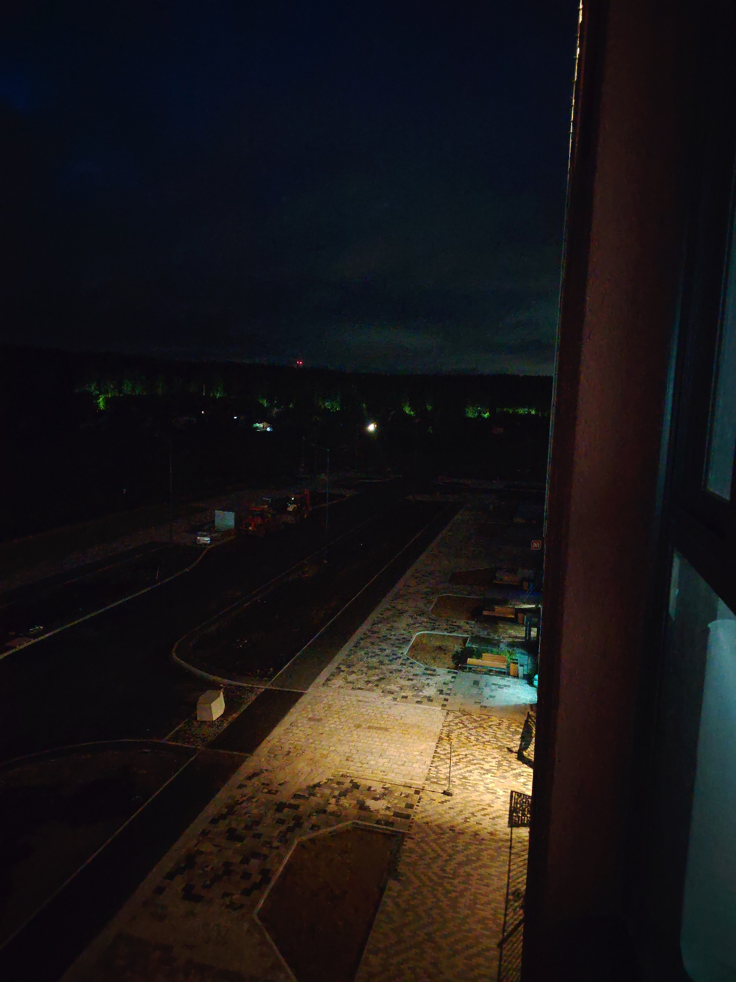 The image is a nighttime aerial view of an under-construction residential area in Yekaterinburg, Russia. In the foreground, a dark, brown, textured building structure is on the right, partially obscuring the view. The ground below is paved with interlocking bricks and some areas of dark soil. Streetlights illuminate sections of the paved area, casting long shadows. Several benches are visible in a landscaped section with some greenery. Further into the scene, a dark asphalt road runs through the development. Construction equipment, including an orange excavator, is present on the road and adjacent areas. In the background, a treeline is illuminated by green lights, suggesting nearby natural or artificial lighting. A red beacon light is visible atop a distant structure. The sky is dark and overcast.