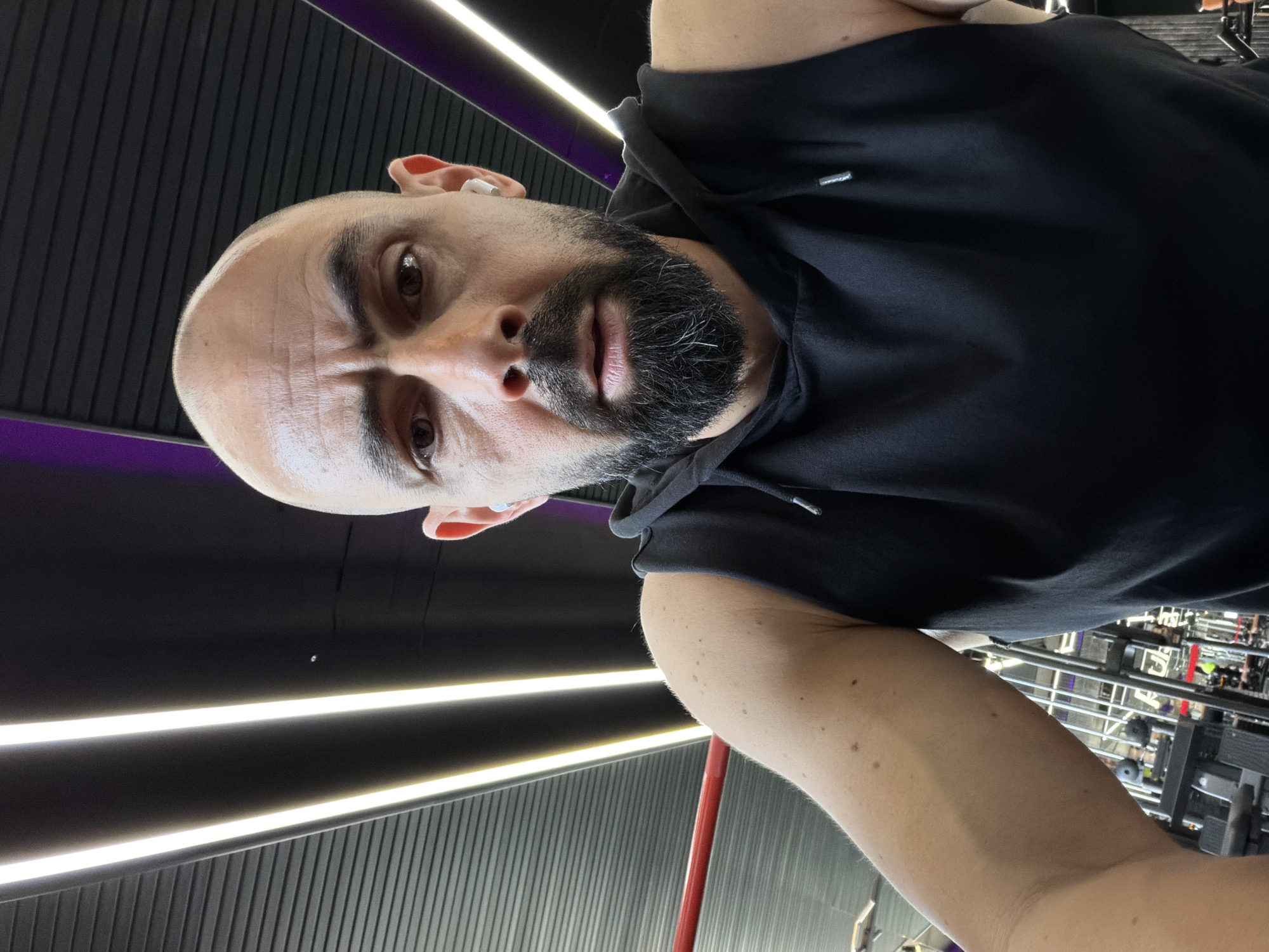 A man with a shaved head and a dark beard is in a gym in Plovdiv, Bulgaria. He is wearing a black hooded sweatshirt. The gym has dark walls with horizontal light strips. Exercise equipment is visible in the background.
