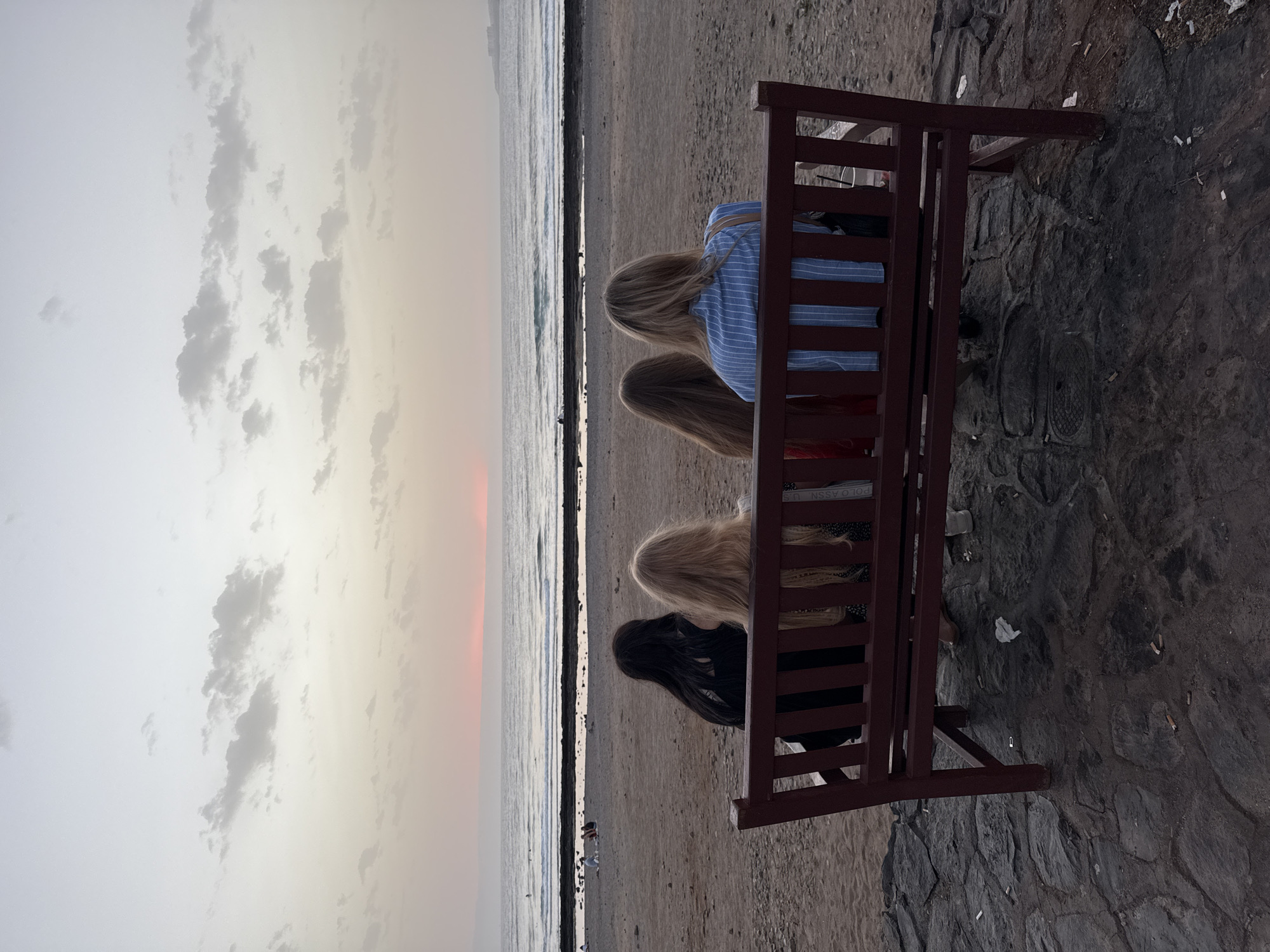 The image depicts four individuals seated on a maroon bench facing a beach and the ocean at sunset. The sky is a gradient of pale yellow and pink, with scattered clouds. The ocean is calm with gentle waves. The beach is sandy with some rocks. In the background, there are a few other people visible on the beach and some buildings on the horizon. The ground in front of the bench is paved with dark stones. This scene is likely located on the coast of Arona, Spain, given the general appearance of the beach and ocean.