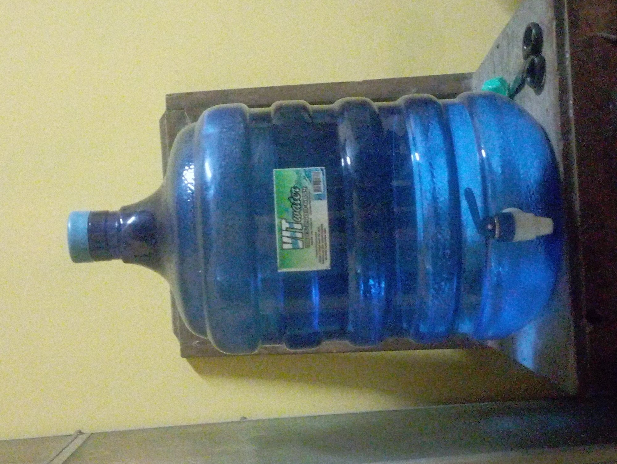 A large blue water jug with a white spigot is placed on a wooden surface in front of a yellow wall. A green and white label with illegible text is attached to the front of the jug. Two black objects, possibly scissors, are next to the jug on the right. The lighting is dim, casting shadows on the objects and the background. The image appears to be taken indoors.
