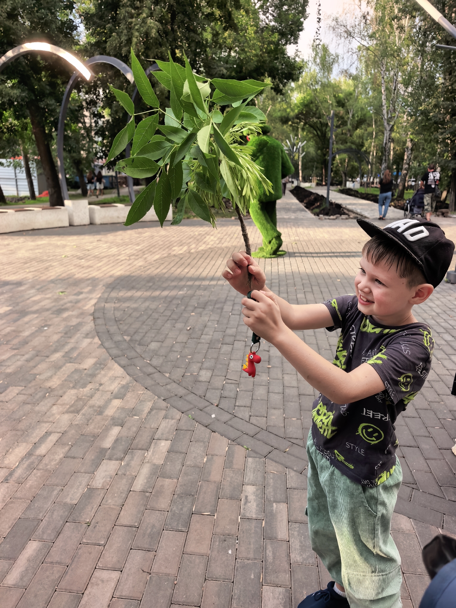In an outdoor urban park, a young boy wearing a black baseball cap with white lettering and a dark t-shirt with neon green accents, corduroy pants, and sneakers smiles as he holds a leafy branch with a small red chicken keychain attached. The boy is standing on a paved walkway with an intricate pattern of interlocking bricks. In the background, a person in a green, furry costume resembling a character is visible. Trees and modern lighting fixtures line the park path, and other park-goers are seen in the distance. The scene is brightly lit by natural light, suggesting it is daytime. This image was taken in the city of Sterlitamak, Russia.