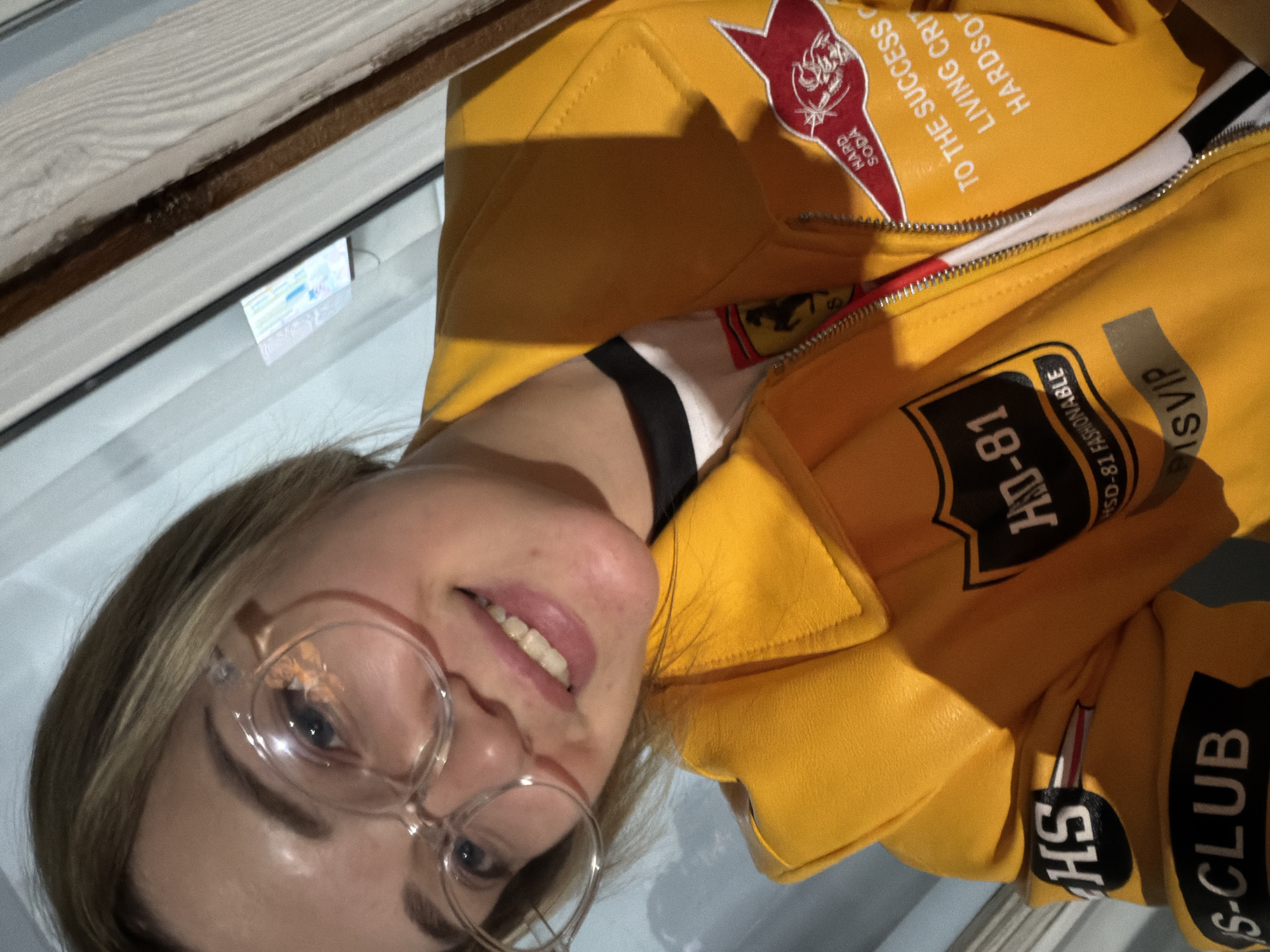 A woman with glasses is wearing a yellow jacket with various patches and lettering on it, including "HARD SODA", "TO THE SUCCESS LIVING CRIT HARDSO", "HD-81", "HSO-81 FASHIONABLE", "SVIP", and "HS-CLUB". The location is indeterminate and no evidence suggests it is in Kazan, Russia.