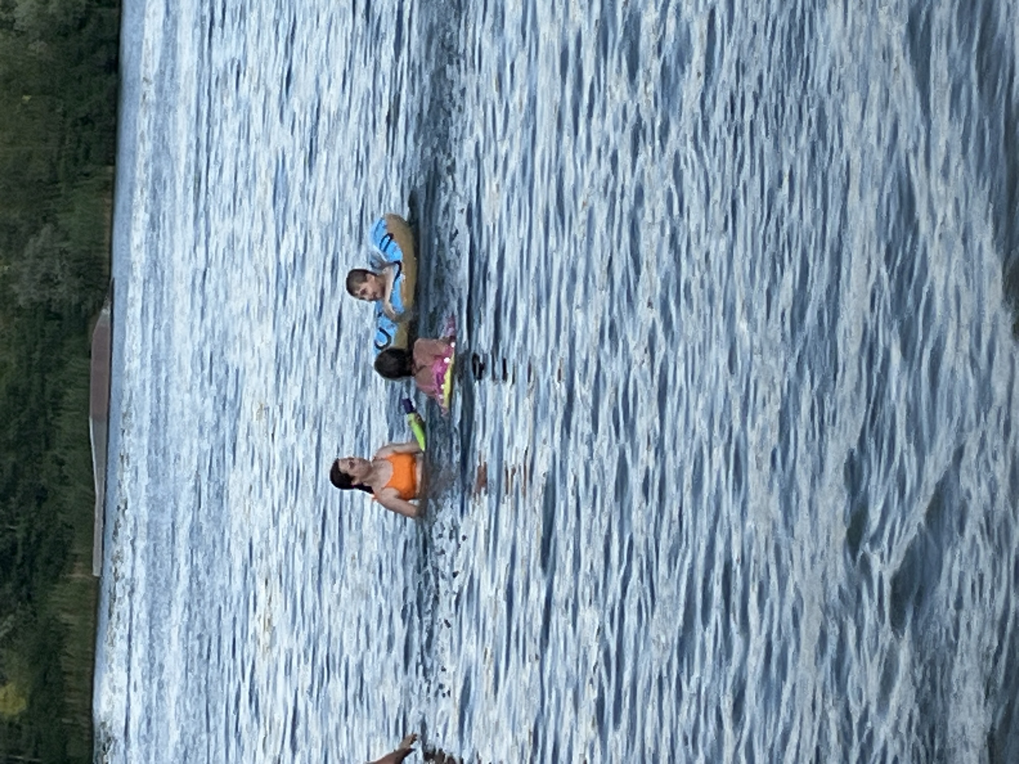 The image shows a group of people enjoying a day in the water. There are at least four individuals visible. One person, wearing an orange top, is floating on their back. Two children are also in the water, one with a blue float and the other with a pink float. Another person appears to be partially submerged in the water. The scene is set on a body of water, likely a lake or a river, given the presence of trees and vegetation along the bank on the left side of the image. The water is rippling, suggesting a gentle breeze. The lighting indicates it is daytime, possibly late afternoon, with soft sunlight creating reflections on the water's surface. The overall mood appears relaxed and recreational. There is no visible text in the image.