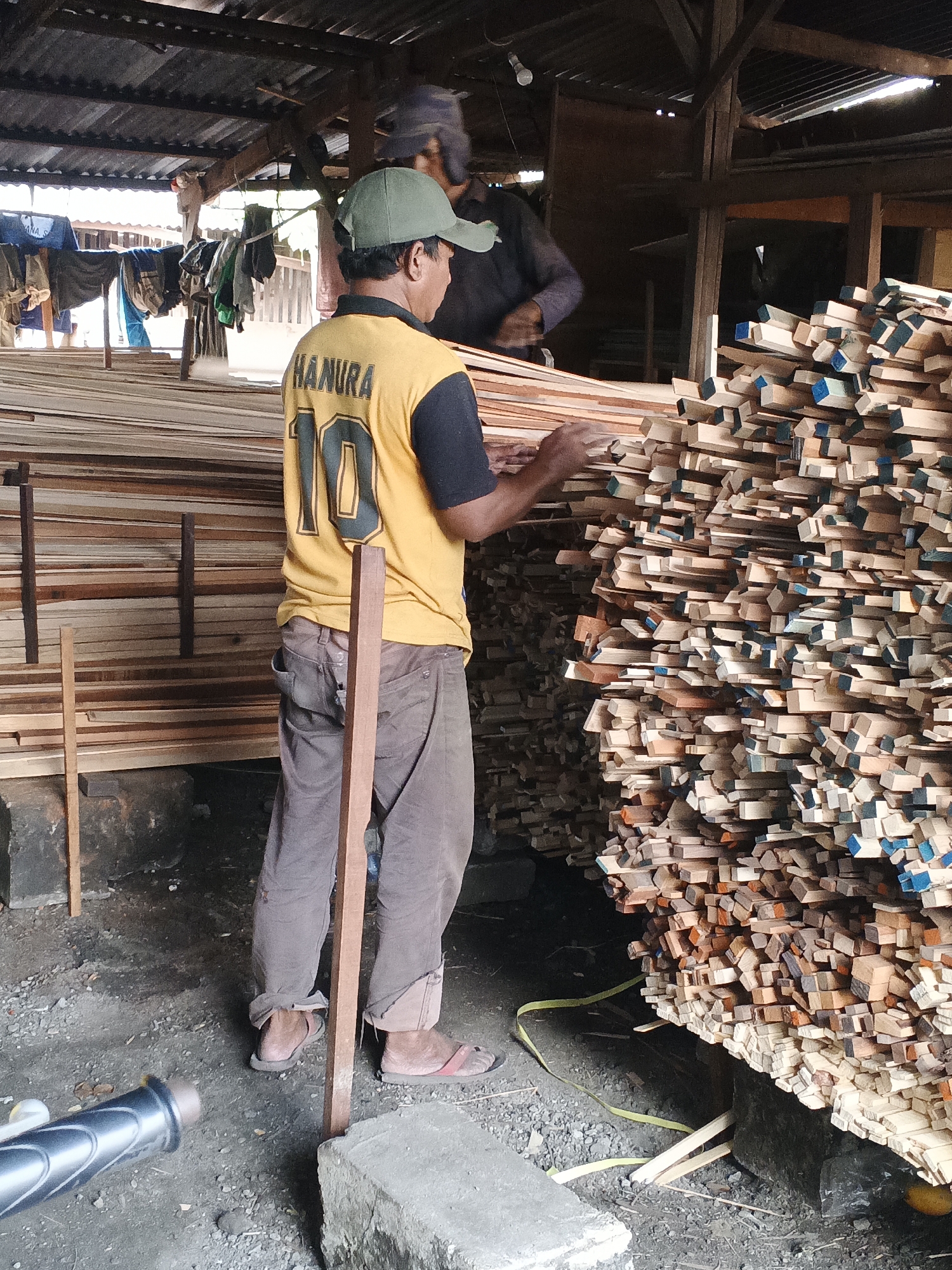 The image captures a scene at a lumber yard in Bakung Pringgodani, Indonesia. In the foreground, a man wearing a green cap and a yellow shirt with "HANURA 10" emblazoned on the back is seen from the back, sorting through stacks of lumber. He is wearing brown pants and sandals. Behind him, another person, partially obscured and wearing a blue cap, is also engaged with the lumber. The environment is an open-air structure with a corrugated metal roof supported by wooden beams. Large quantities of sawn lumber are piled high, neatly stacked and forming significant walls of wood. The ground is covered with dirt and gravel. To the left of the image, laundry hangs on a clothesline. A portion of a bicycle handlebar is visible in the lower left corner. The overall impression is one of industry and manual labor within a rural or semi-rural setting.