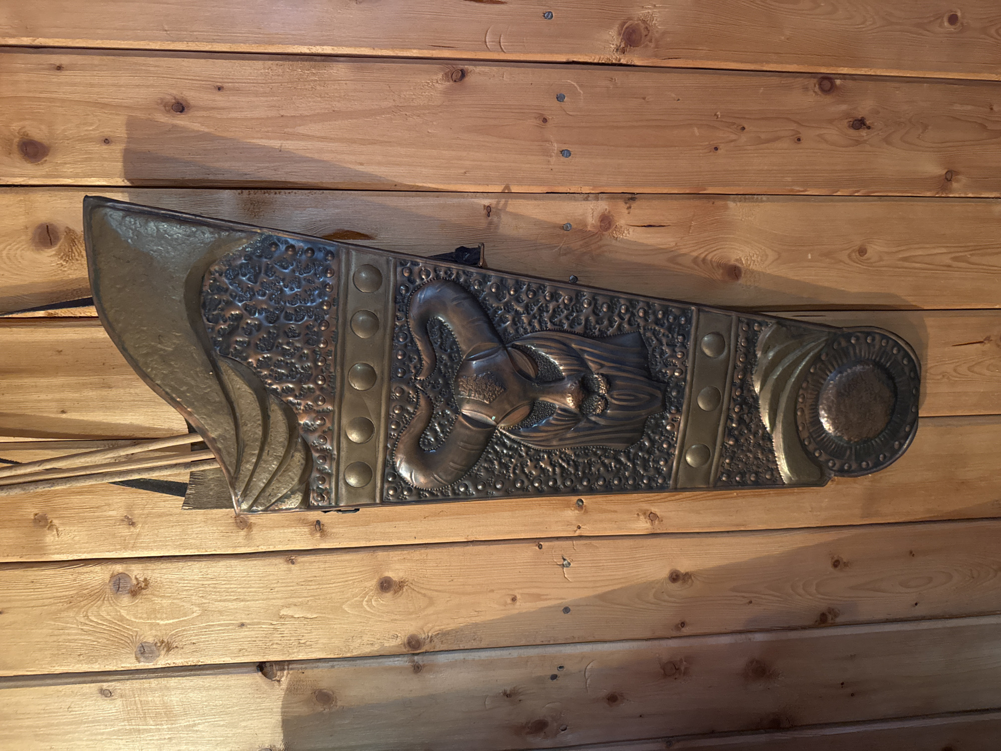 The image shows a decorative, ornate metal object mounted on a wooden wall made of light-colored planks. The metal object is roughly triangular in shape with various sections featuring different textures and designs. One prominent design element appears to be an elephant head with its trunk curved upward. A small number of arrows or similar objects are visible extending from the left side of the metal object. The setting and city are unknown.