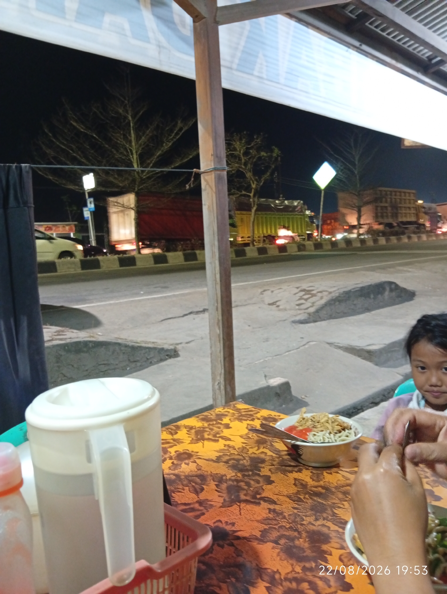 This is a nighttime, slightly low-light, eye-level outdoor shot taken at street level in the city of Sengeti, Indonesia. In the foreground, a table is set with food, including a bowl of noodles with chili sauce and fried shallots, and a red plastic basket. To the right, a person's hands are visible, reaching towards the food, and a child's face is partially visible looking towards the camera. Behind the table, a white pitcher is prominent. The background depicts a street scene with traffic and buildings. A car and several trucks are on the road, illuminated by streetlights and their own headlights. Trees, some bare, are visible against the dark sky, and a diamond-shaped sign is illuminated with a yellowish light. The overall impression is of a casual dining setting along a busy road at night.