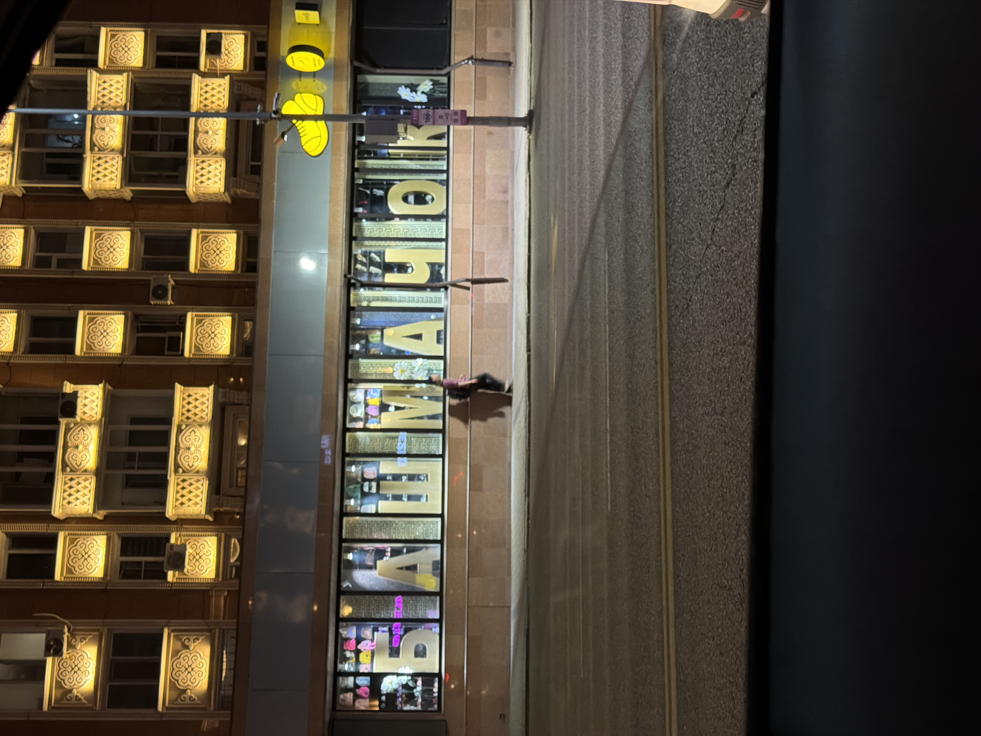 A nighttime exterior street view shows a building in Astana, Kazakhstan. The building has multiple floors with illuminated decorative elements around the windows and balconies. Below, a storefront displays large, golden Cyrillic letters spelling out "БАШМАЧОК" (Bashmachok). Inside the store windows, various bags and shoes are visible. A person is walking in front of the store, casting a shadow on the sidewalk. Across the street, a road with yellow lane markings is visible, and a portion of a vehicle is parked on the right side of the frame. A street lamp and a sign with Cyrillic text and the number "100+" are also present.