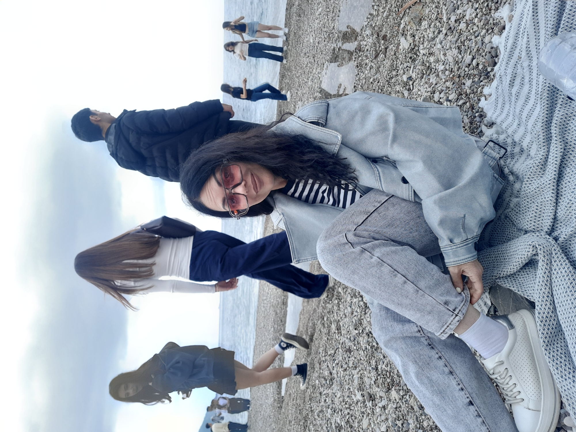 A portrait-oriented photograph, rotated clockwise, captures a group of people enjoying a pebbly beach in Kumluca, Turkey, under an overcast sky.
In the foreground, a woman with long dark hair and pink-tinted sunglasses is seated on a light-colored, patterned blanket. She is dressed in a striped top, a light blue denim jacket, light-wash jeans, white socks, and white sneakers, and is looking towards the viewer with a subtle smile. A clear plastic water bottle rests on the blanket beside her.
Further back on the beach, several other individuals are visible. To the left of the main subject, a man in a black puffer jacket stands, looking out towards the water. Behind him, another woman with long brown hair, wearing a white long-sleeved top and dark pants, is seen from behind, walking near the water's edge. Additional figures, some appearing as reflections or silhouettes, are scattered along the calm sea and pebbled shore in the mid-ground and background. One person wearing a dark top and black sneakers is also partially visible closer to the water.
The scene depicts a relaxed daytime outing on a tranquil pebbly beach, likely along the Mediterranean Sea, characterized by a soft, diffused light from the overcast sky. There is no visible text in the image.