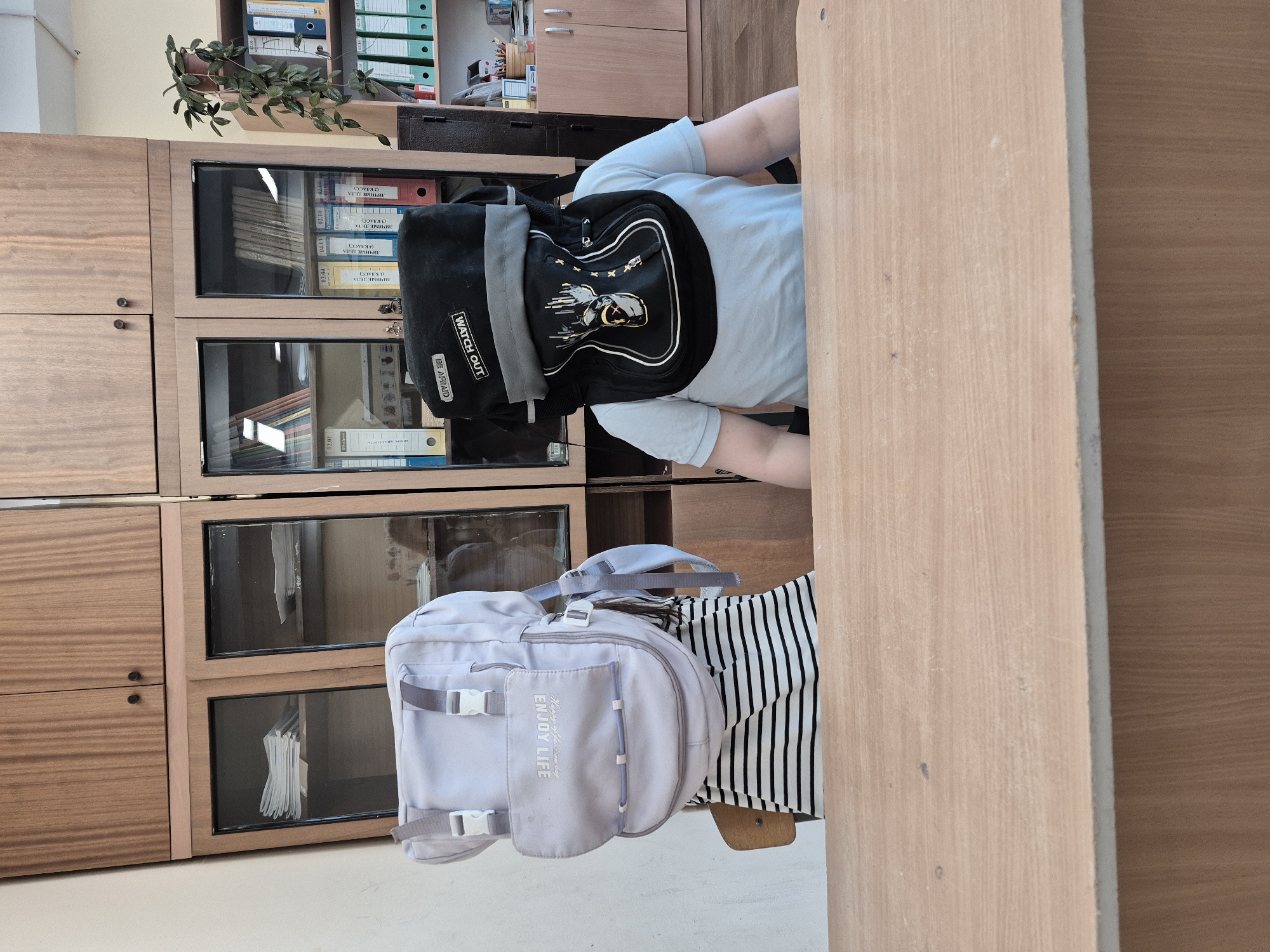 Two individuals are seated at a wooden desk in what appears to be a classroom or office setting in Chaikovsky, Russia. Both are viewed from behind, facing away from the camera. The person in the foreground wears a light blue t-shirt and a black backpack, while the person further back wears a black and white striped t-shirt and a light grey backpack.
The setting includes a large wooden cabinet unit with both solid doors and glass-fronted sections, filled with binders and books. A small potted plant with green leaves is placed on top of the cabinet. The room is well-lit, suggesting daytime.
Notable details include the text on the backpacks and binders. On the black backpack, the phrases "WATCH OUT" and "BE AFRAID" are visible on a black and white patch. The grey backpack features the text "ENJOY LIFE" along with other decorative, stylized writing. The binders within the cabinet are labeled in Cyrillic, with "ЛИЧНЫЕ ДЕЛА" (Personal Files) prominently displayed, along with class numbers such as "(1 КЛАСС)" (Class 1), "(3 КЛАСС)" (Class 3), and "(4 КЛАСС)" (Class 4), and numerical codes like "02.01," "02.08," "02.10," "04.03," and "03.04." These labels suggest an educational or administrative environment.